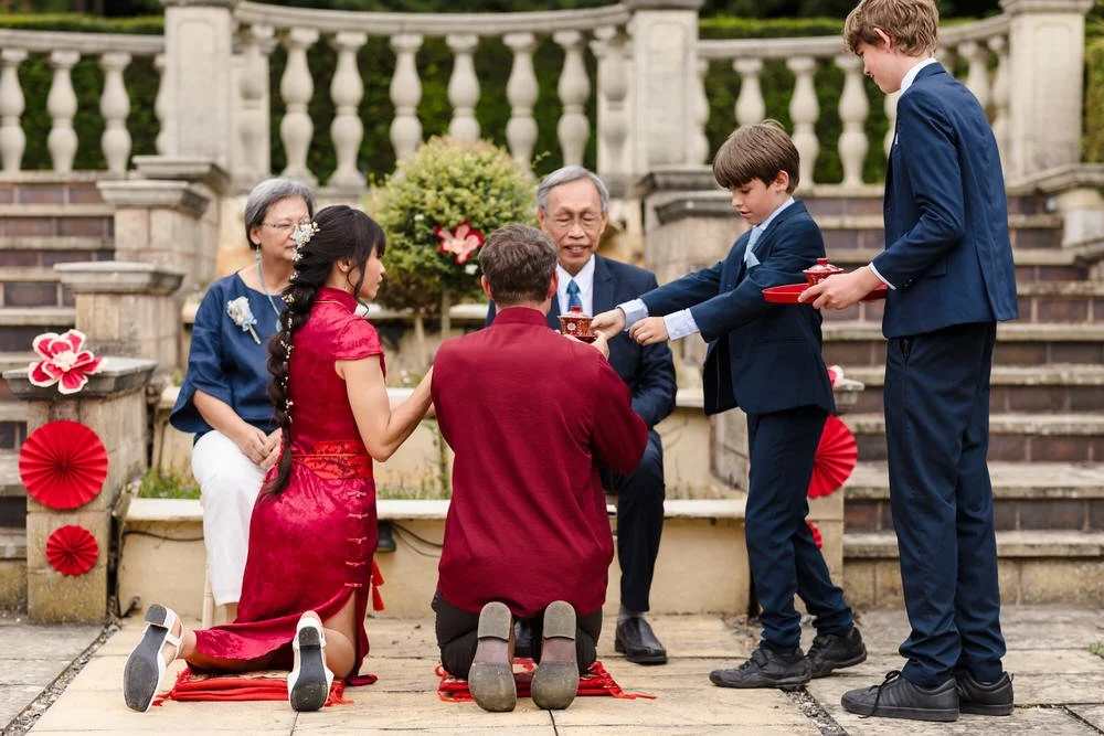 Christine and Greg kneeling in traditional red attire, offering tea to Christine's parents on The Welcombe Hotel terrace.