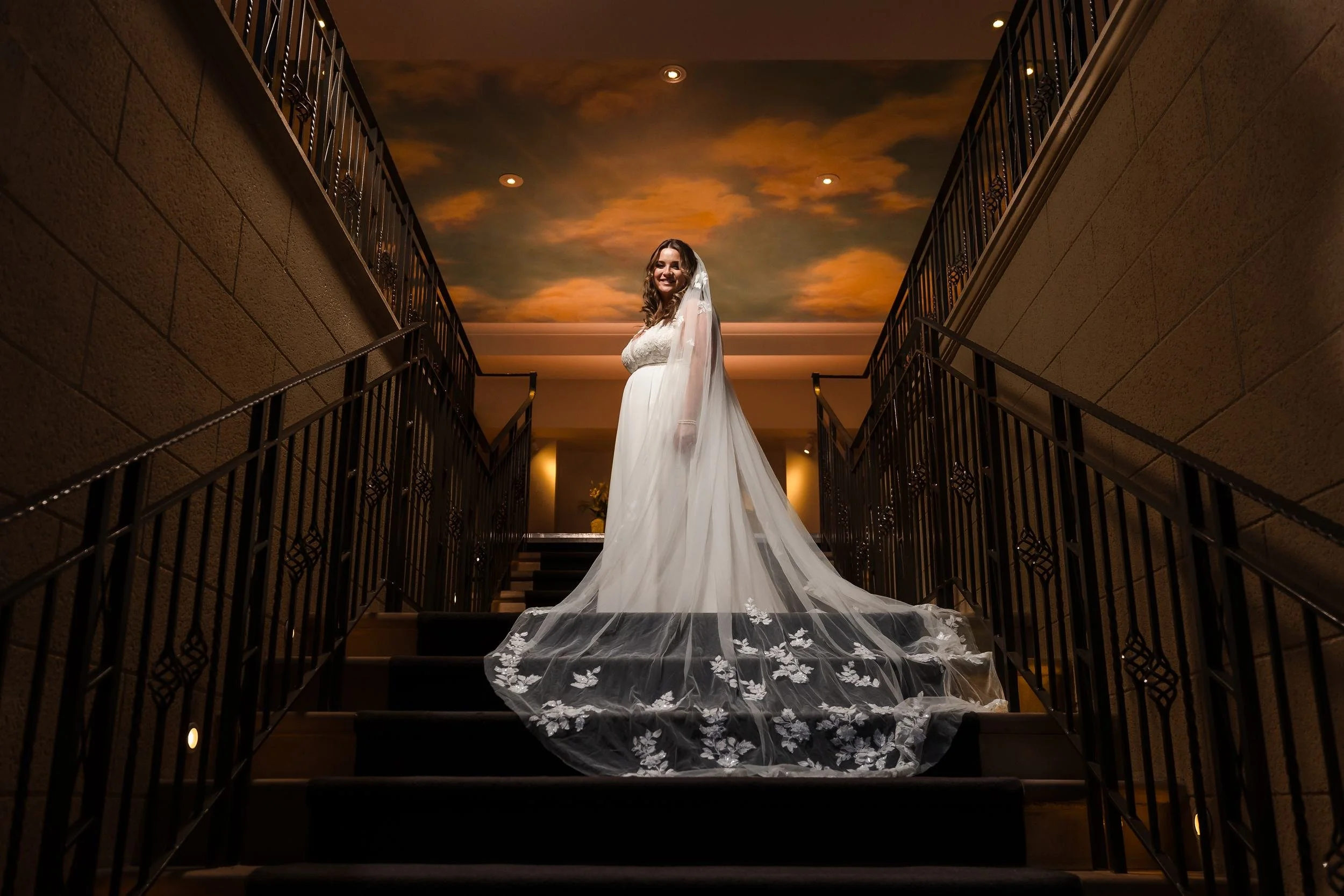 Holly standing alone on the staircase at Nailcote Hall with her embroidered veil spread down the steps, lit by off-camera flash beneath a warm painted-sky ceiling