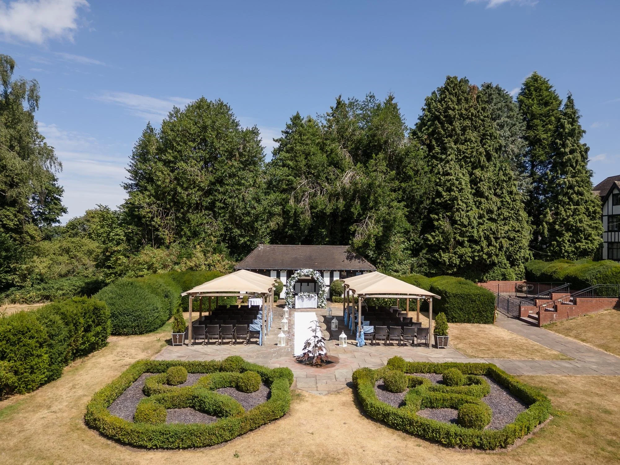 Aerial view of the outdoor ceremony area at Hogarths Stone Manor, set up with a white aisle, blue and white floral arch and two canopied seating areas behind the knot garden