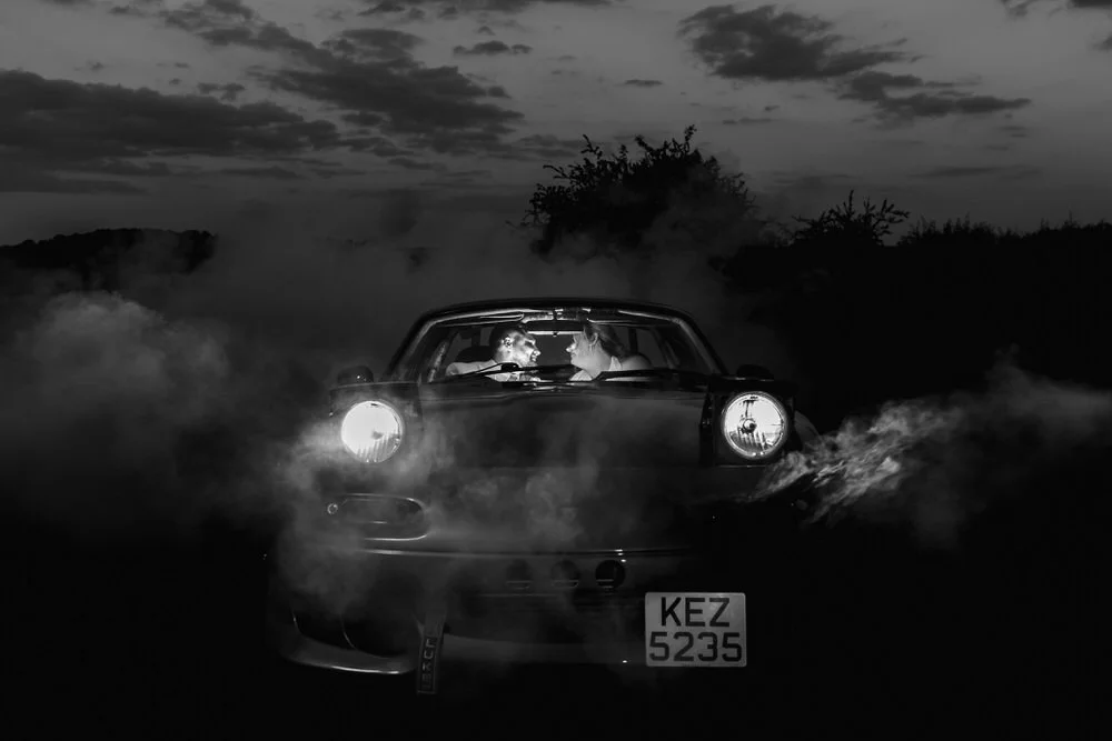 Dramatic black and white wedding portrait of bride and groom in sports car with headlights illuminated surrounded by atmospheric smoke effects at dusk.