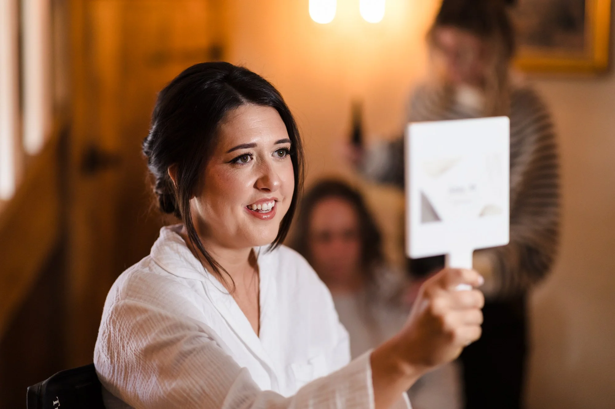 Bride in a white robe holding up a mirror during morning preparations, candid bridal prep photography