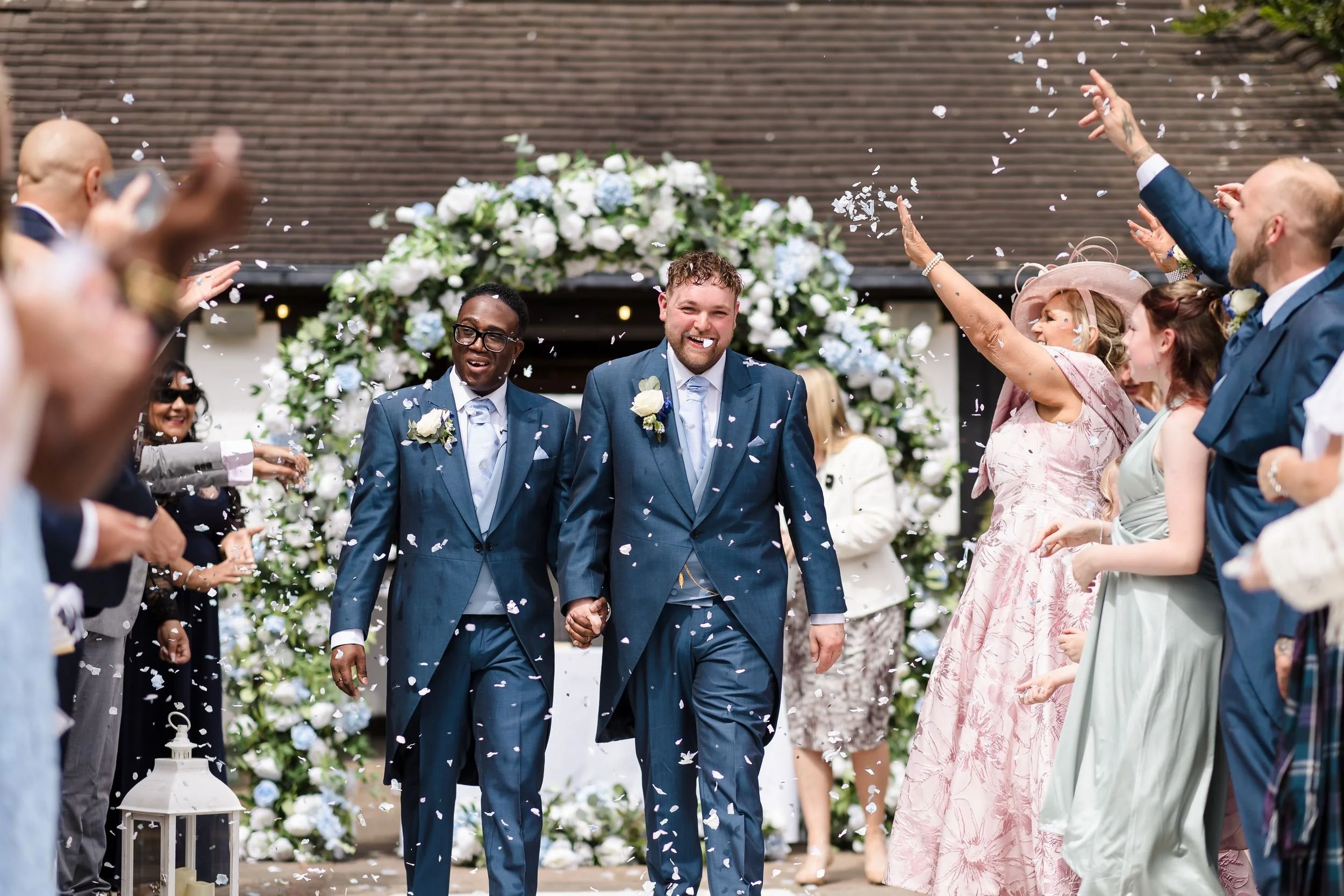 Liam and Shaun walking hand in hand through a shower of white confetti from their guests after the ceremony at Hogarths Stone Manor