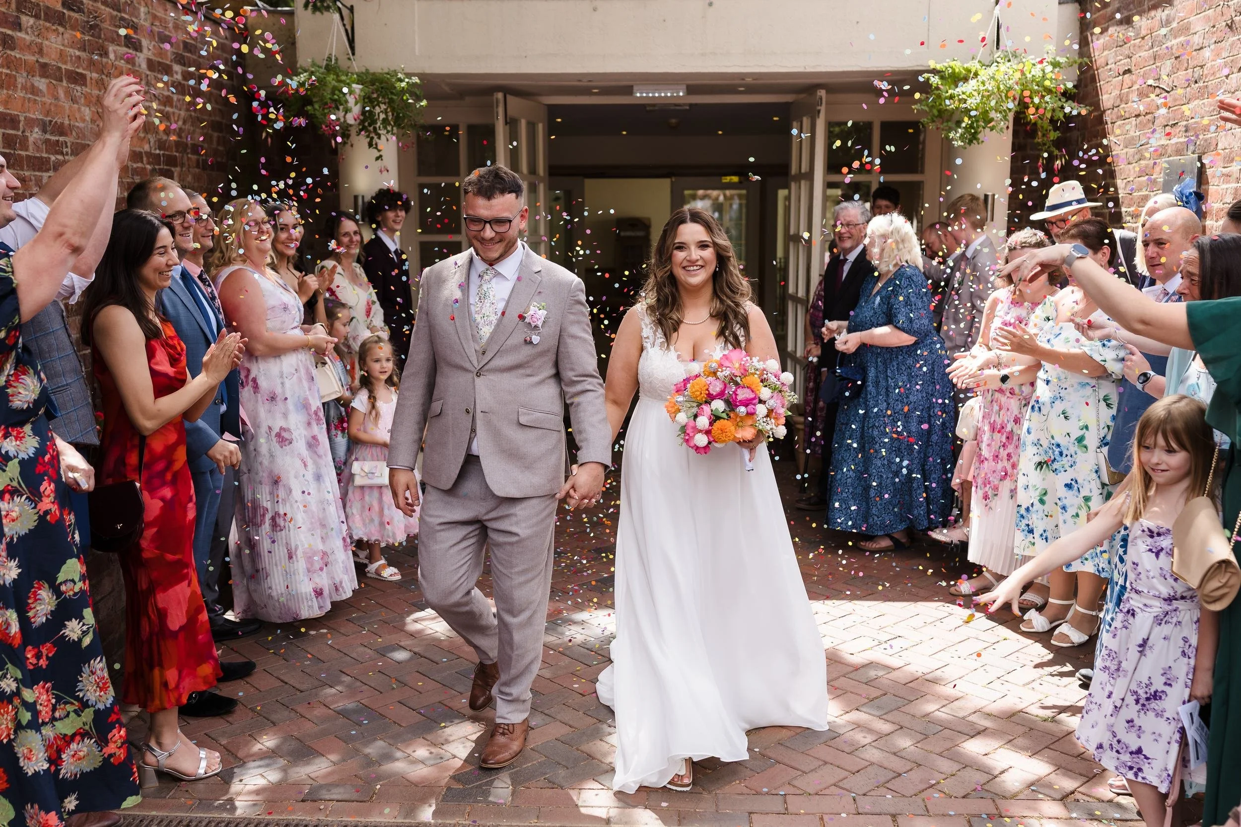 Holly and Dan walking hand in hand through colourful confetti thrown by two lines of guests outside Nailcote Hall