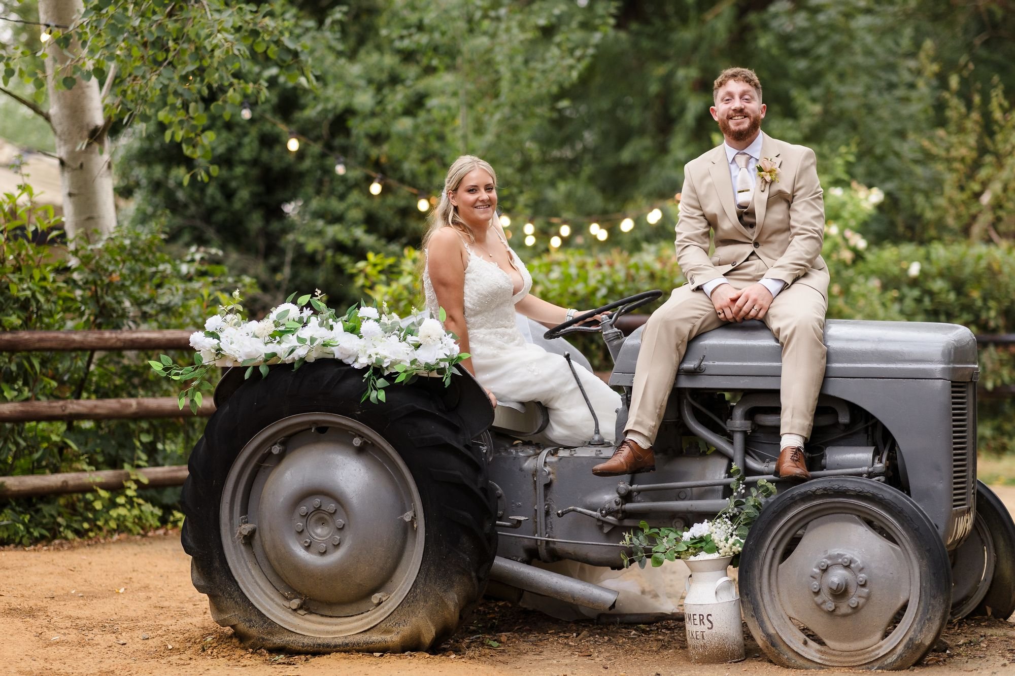 Abbie and Sean sitting on a grey vintage tractor decorated with white roses, festoon lights behind, at Bennetts Willow Barn