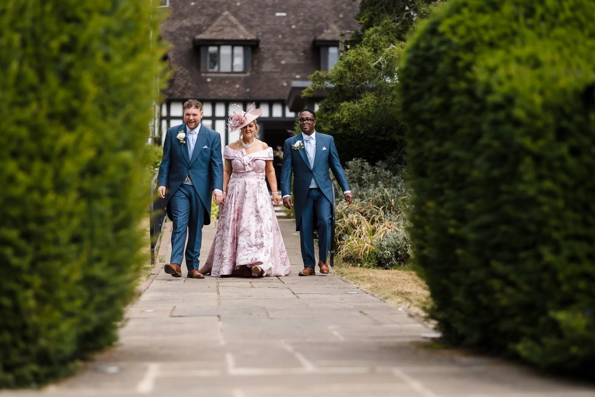 Liam and Shaun walking along the hedge-lined path towards the outdoor ceremony with a guest in a pink floral gown and fascinator
