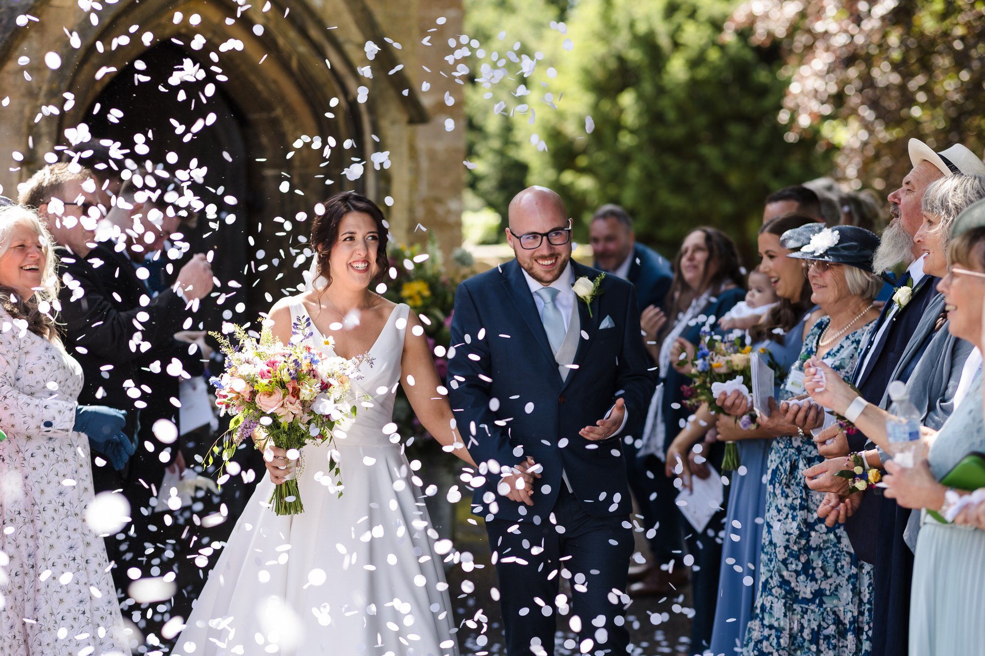 Bride and groom laughing under a shower of confetti outside St Leonard's Church, candid wedding photography Worcestershire