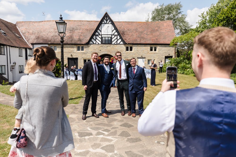 Guests take a smartphone photo of the groomsmen outside the barn at Karma Salford Hall.