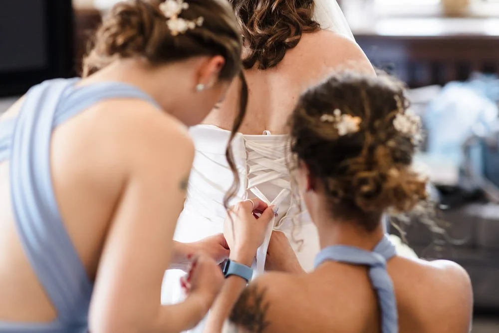Bridesmaids fastening the lace-up back of the bride’s dress during preparations at Karma Salford Hall.