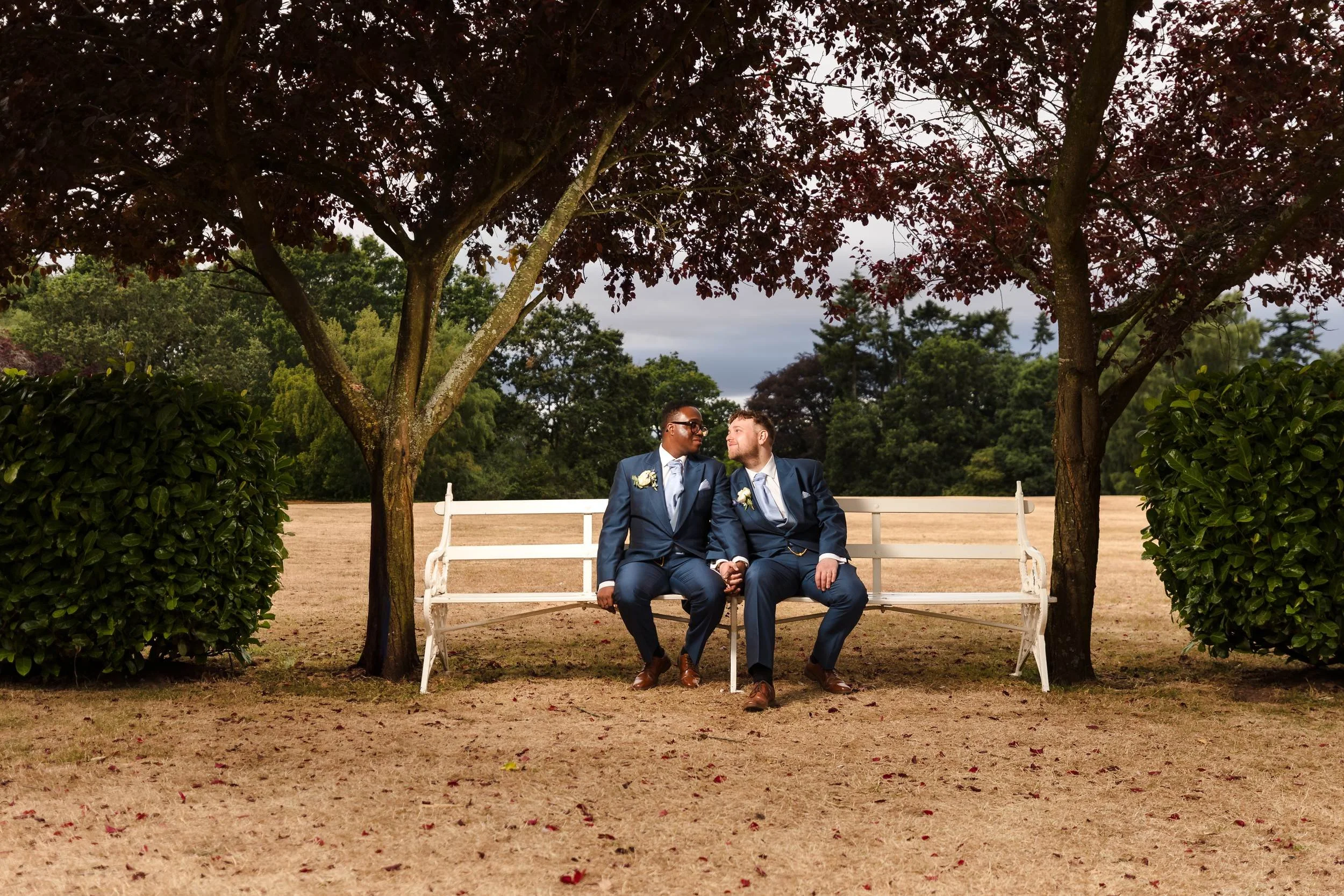 Liam and Shaun sitting close together on a long white bench between two red-leaved trees on the lawn at Hogarths Stone Manor, under a dramatic evening sky