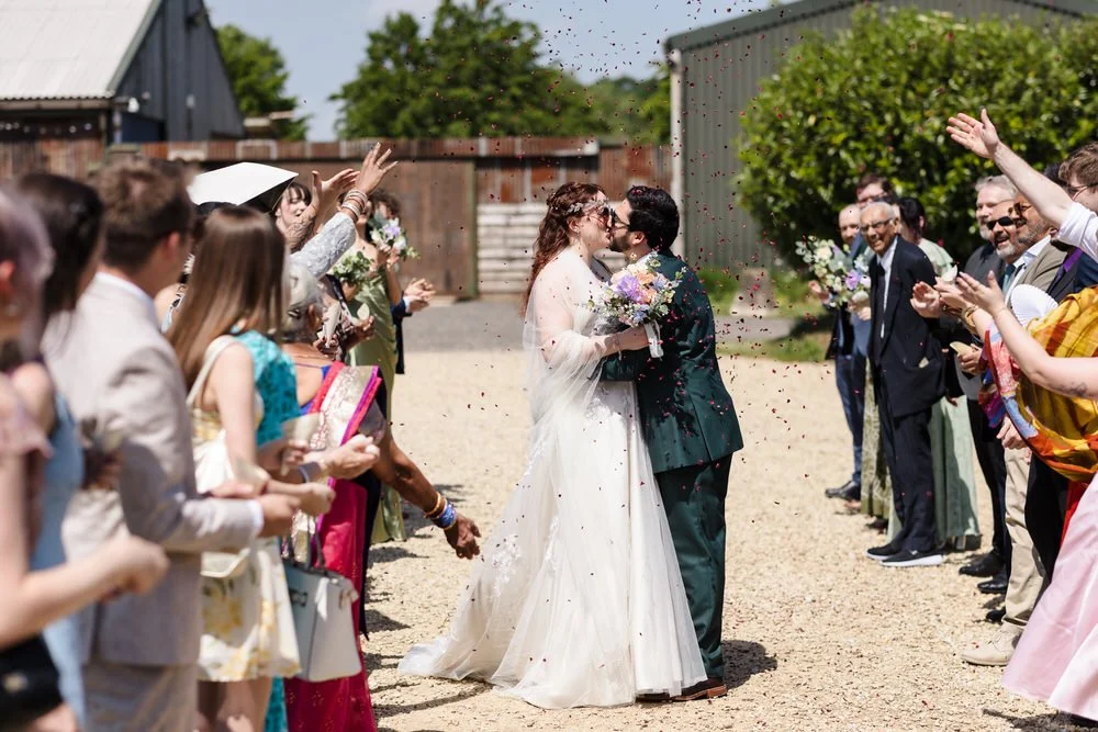Bride and groom kissing under confetti as guests celebrate outside the barn at Huntsmill Farm.