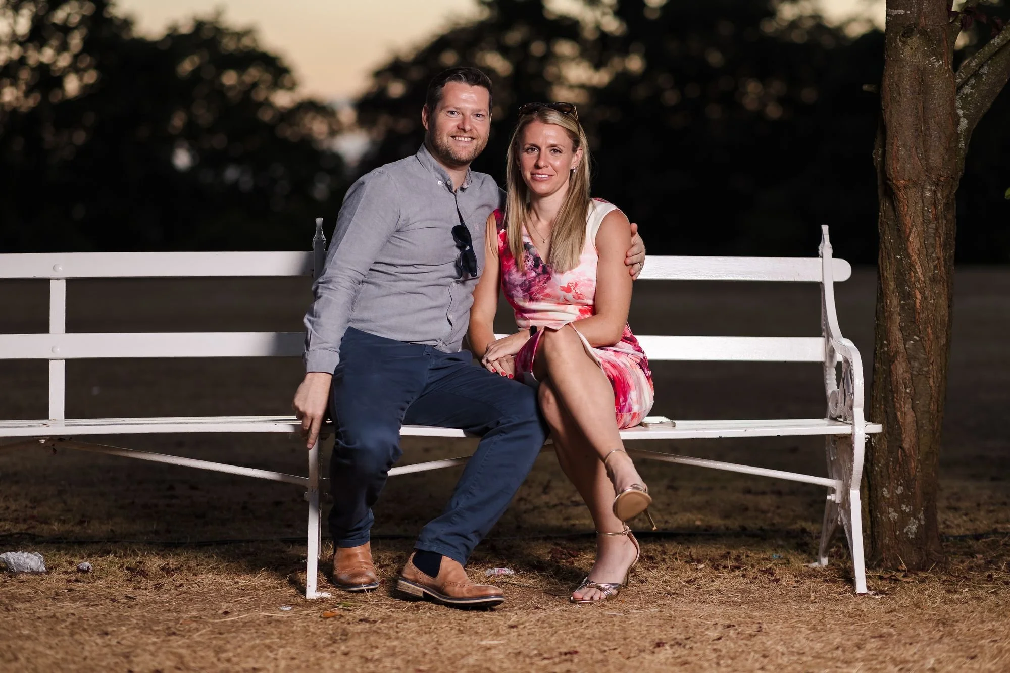 Two wedding guests sitting together on a white garden bench at sunset, lit with flash, at Hogarths Stone Manor