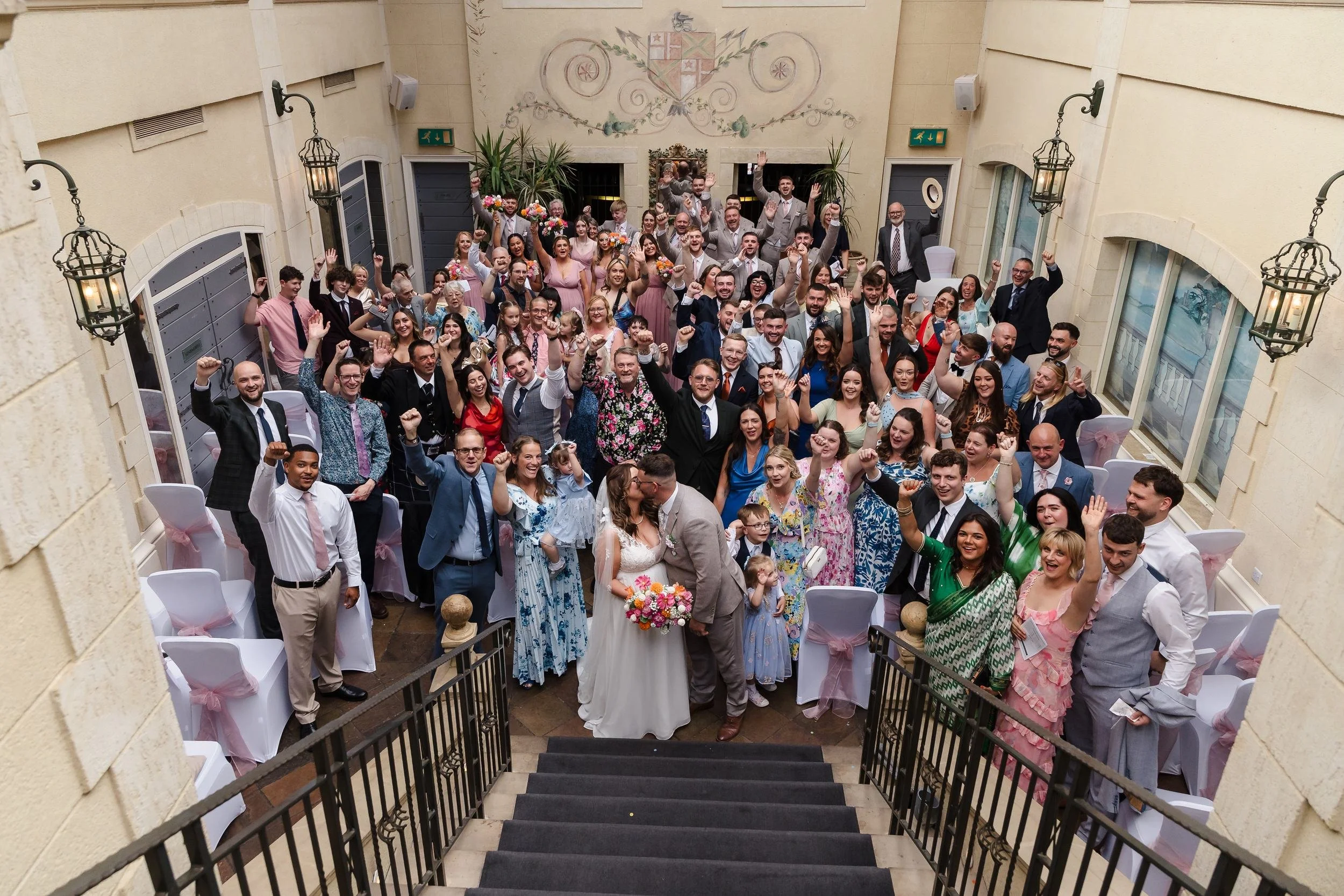 Holly and Dan kissing at the foot of the staircase, surrounded by all their cheering wedding guests in the ceremony room at Nailcote Hall