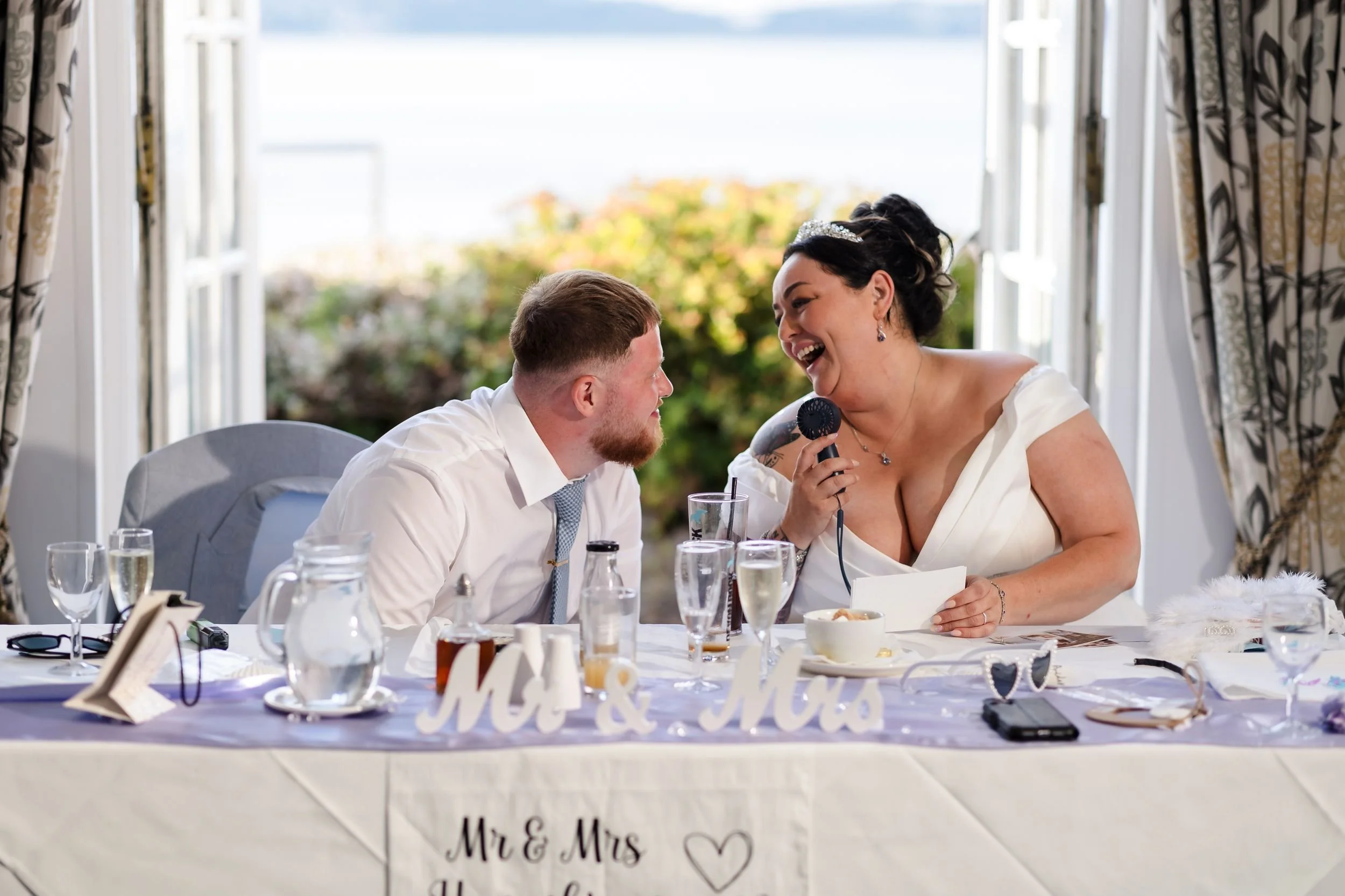 Bride and groom laugh at their sweetheart table during the speeches, with the bay visible through the open doors