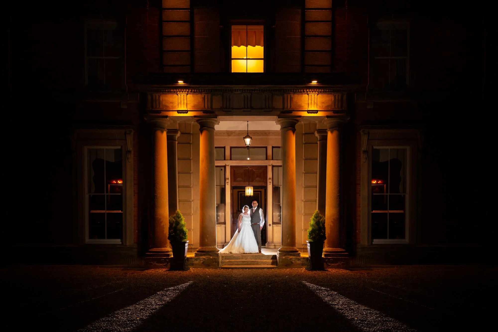 A bride and groom standing on the front steps of a large, illuminated house at night. The house has tall columns and a warm yellow light, with two potted plants flanking the entrance.
