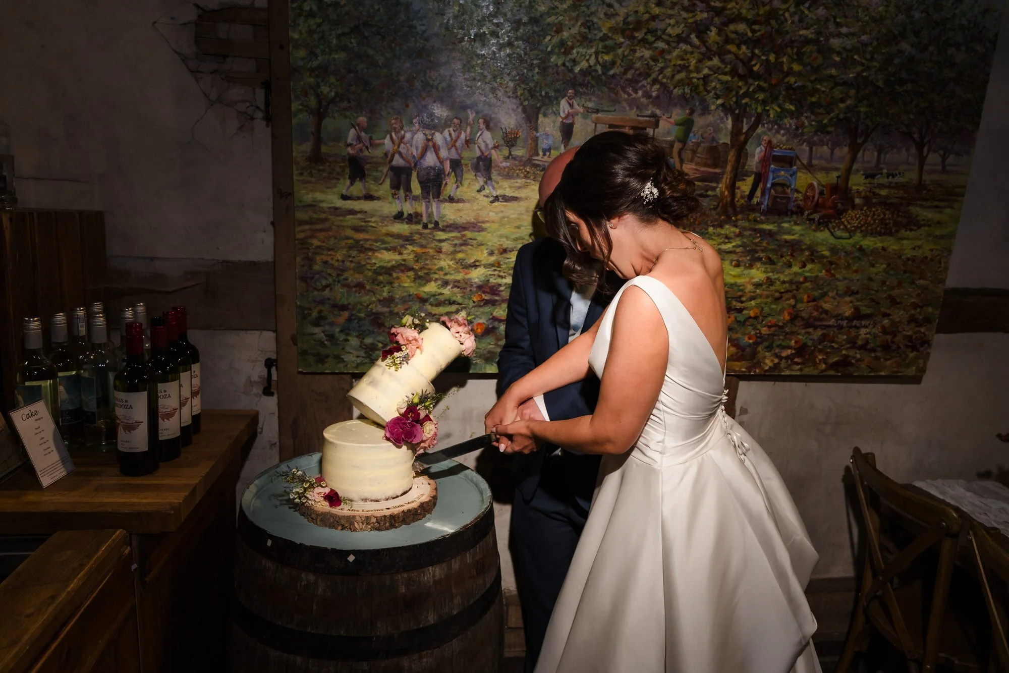 Bride and groom cutting a three-tier wedding cake in front of an orchard mural at The Fleece Inn, Worcestershire