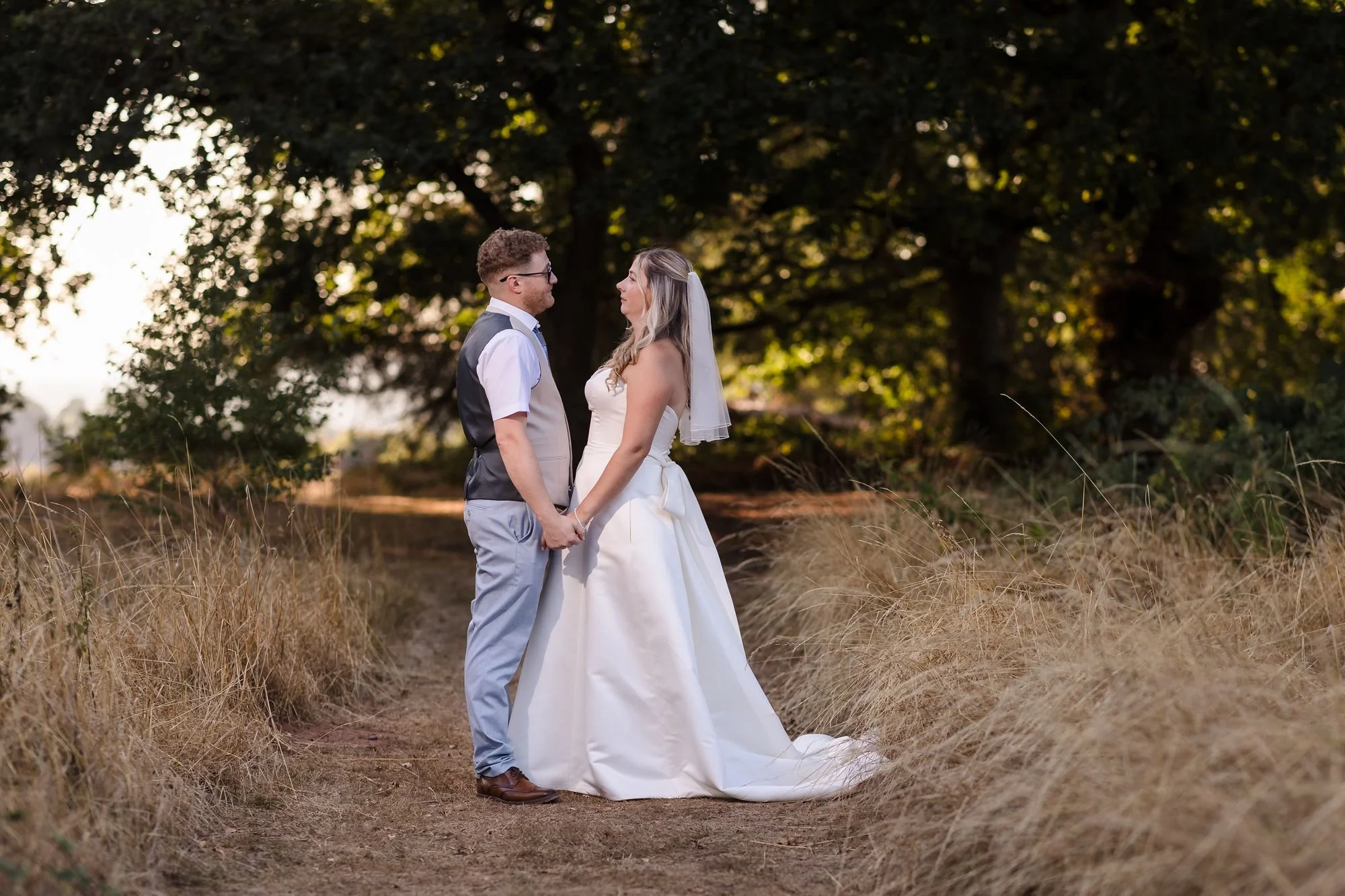 Cheryl and Josh holding hands on a path between sun-dried grasses beneath the trees at the far end of the grounds