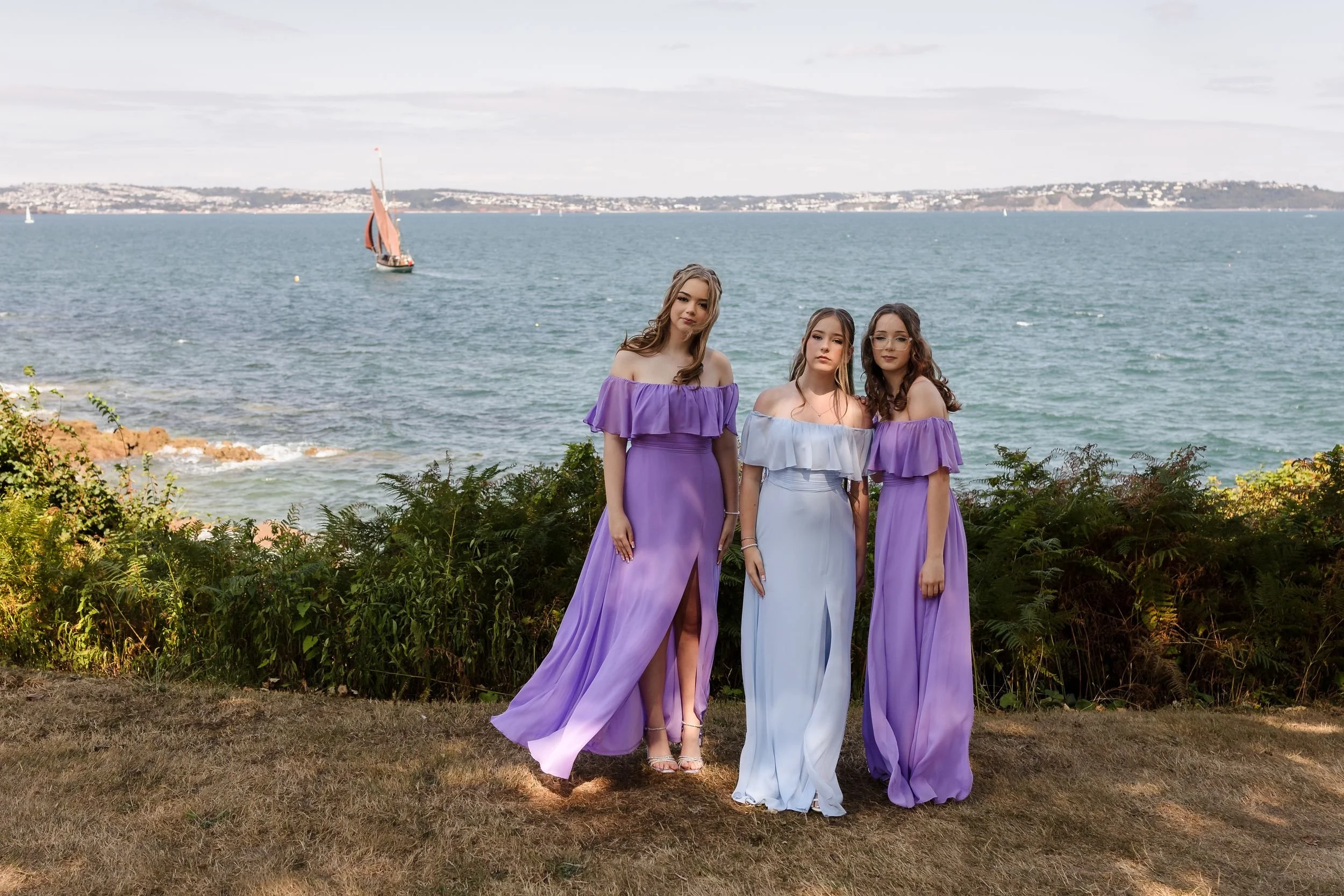 Three bridesmaids in lilac and pale blue dresses on the lawn at Berry Head Hotel, with the bay behind them