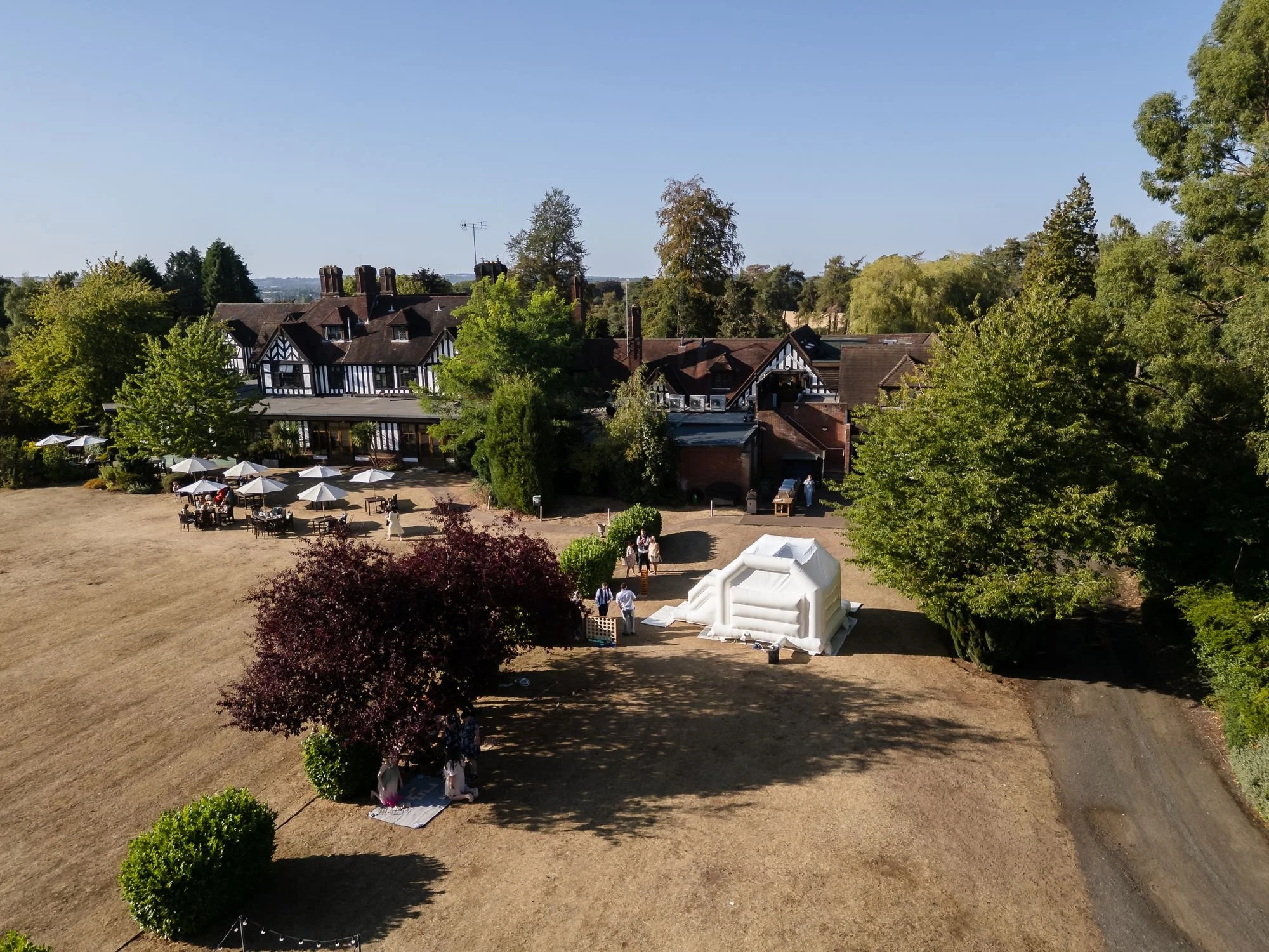 Aerial view across the sunlit lawn at Hogarths Stone Manor with guests, parasols, a red-leaved tree and a white bouncy castle