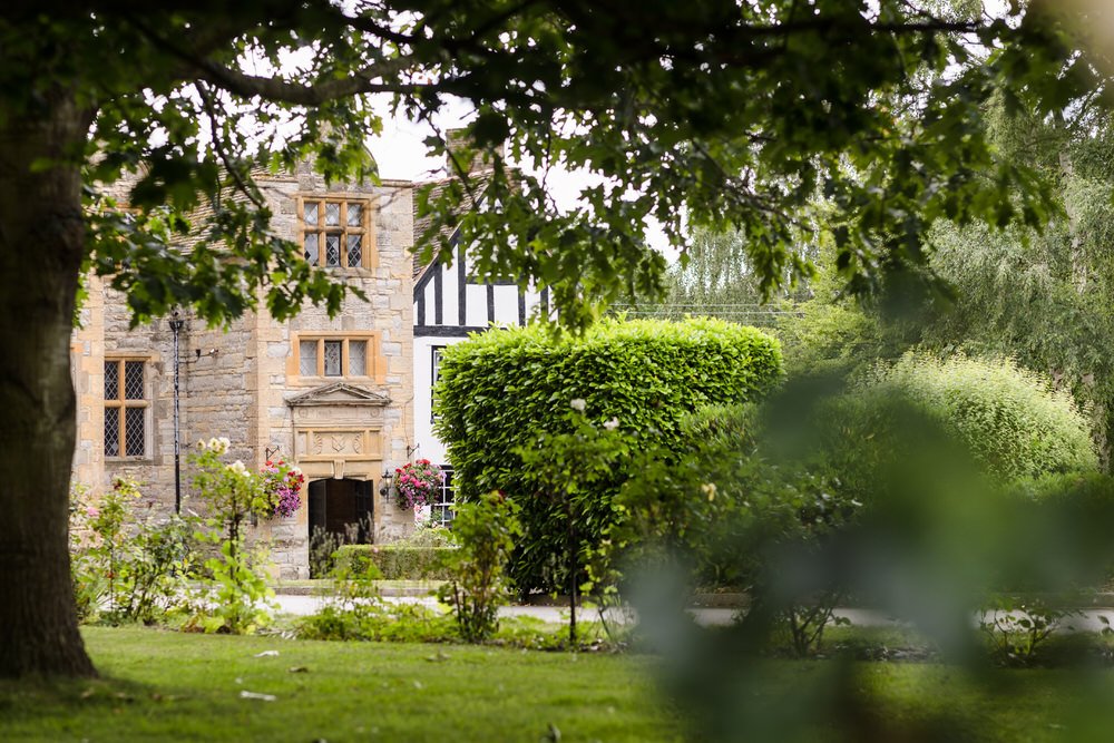 Historic stone entrance of Karma Salford Hall framed by lush summer greenery and hanging baskets.