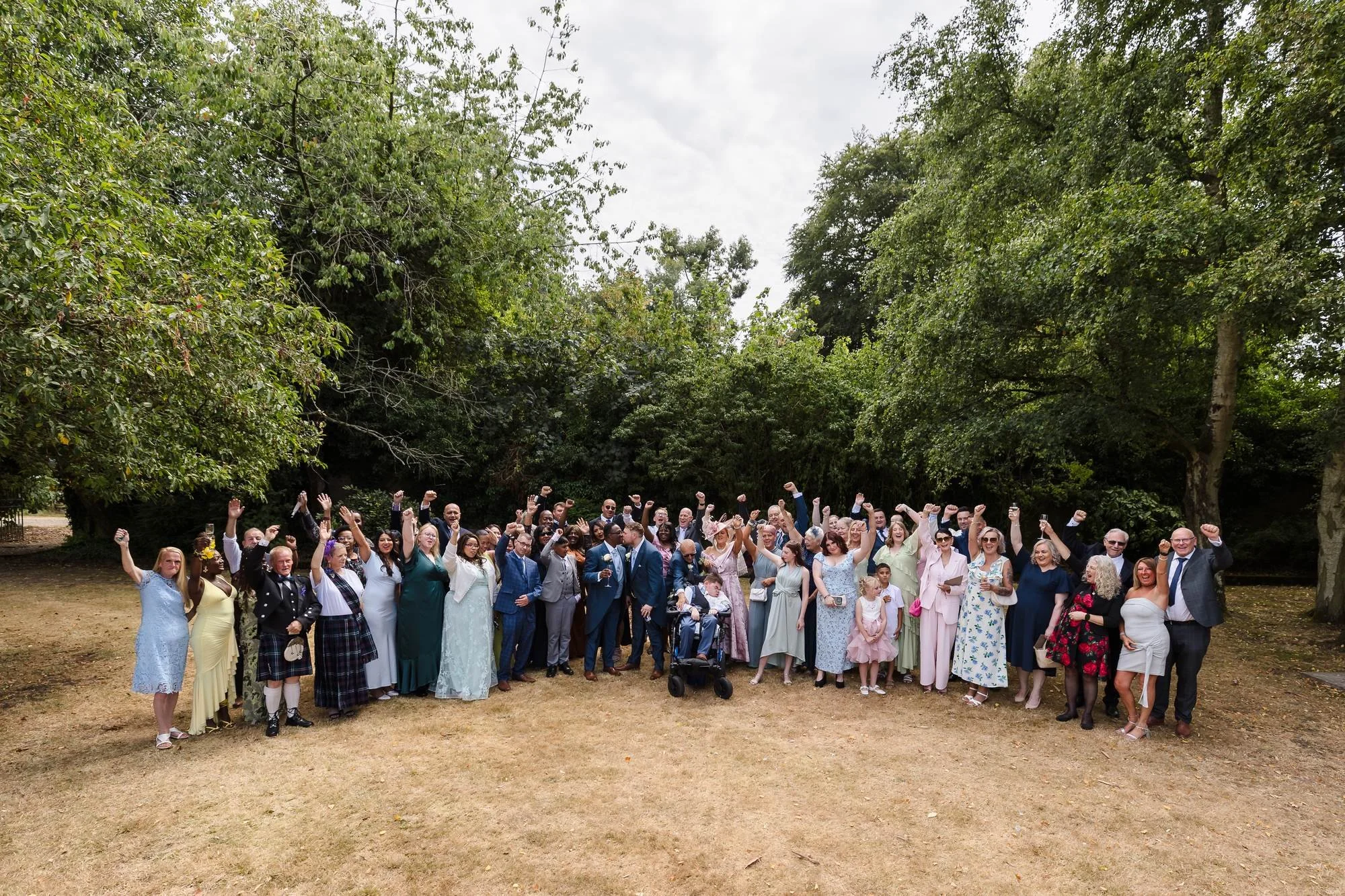 Large group photograph of Liam and Shaun with all their wedding guests, arms raised and cheering among the trees at Hogarths Stone Manor