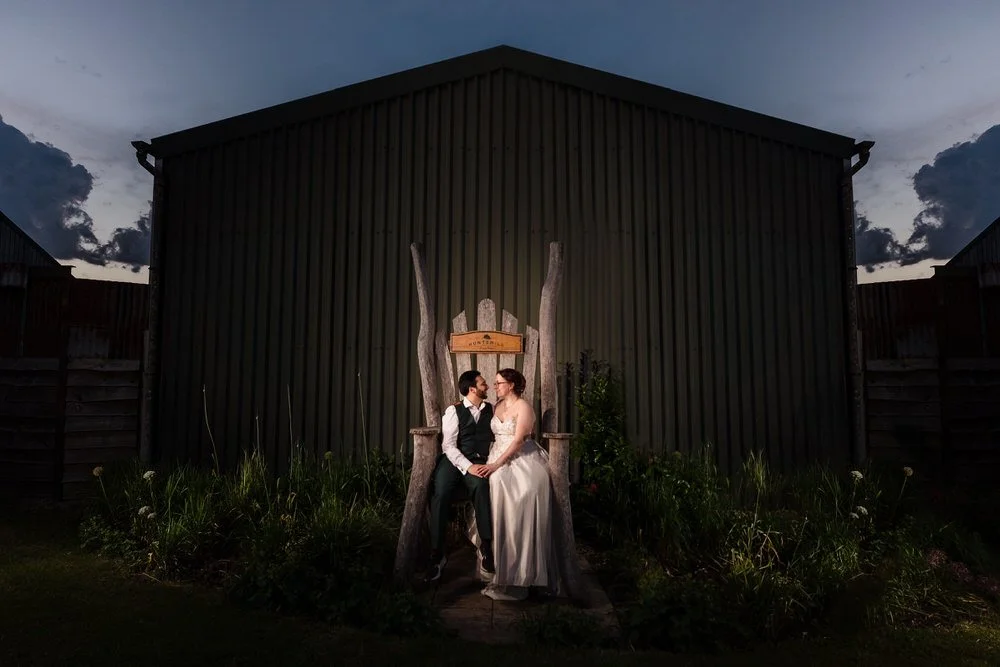 Bride and groom sitting in the giant wooden chair at Huntsmill Farm at dusk.