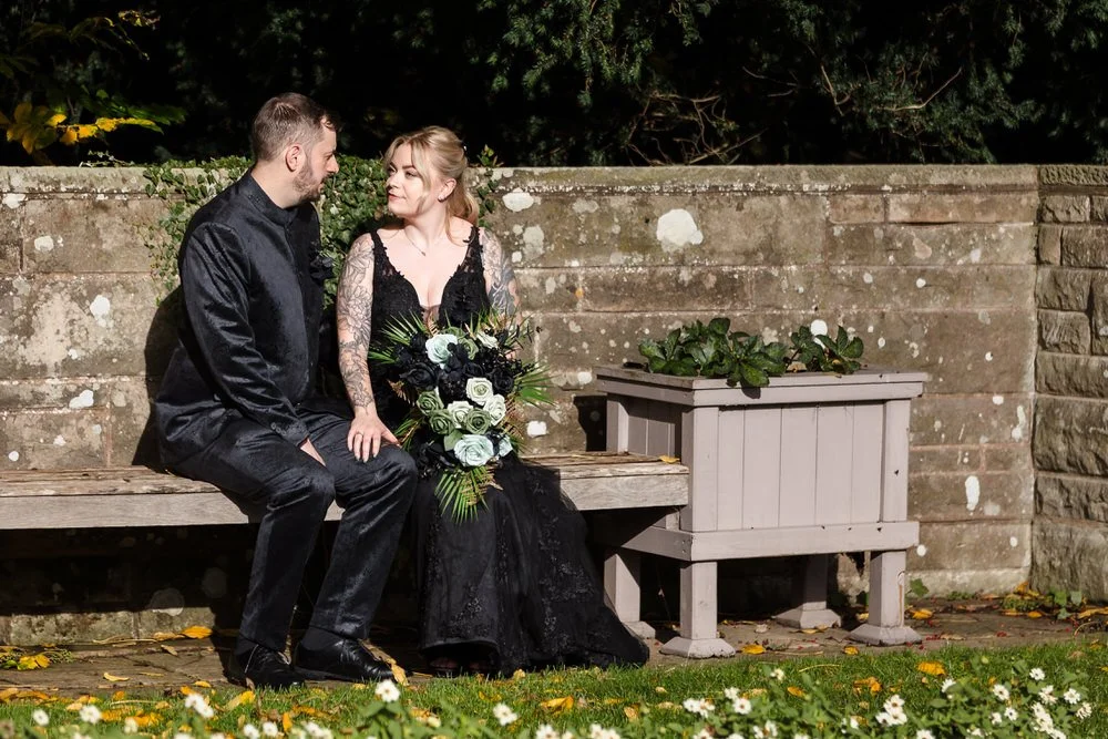 Gothic wedding couple relaxing on garden bench at Arley House with historic stone wall backdrop, natural moment photography Worcestershire