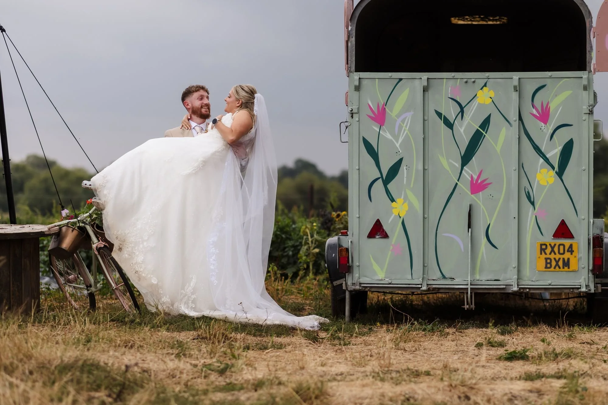 Sean lifting Abbie beside a horsebox painted with flowers and an old bicycle on the farm at Bennetts Willow Barn