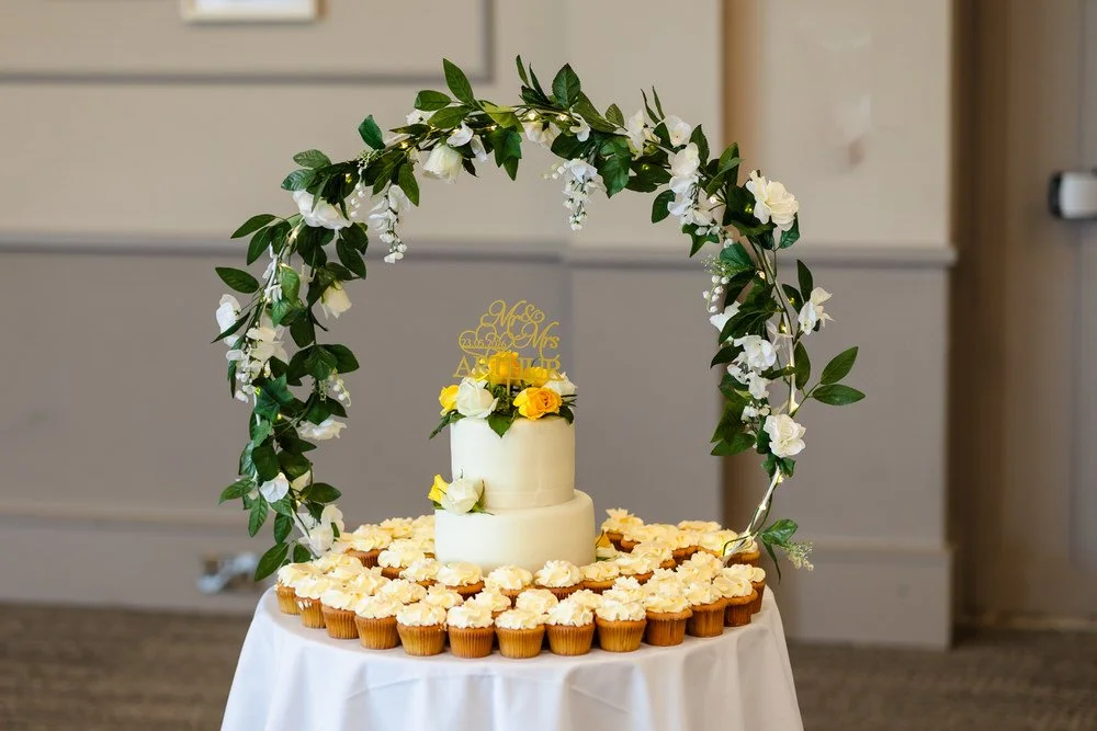Two-tier wedding cake with floral arch and cupcakes at a De Vere Cotswold Water Park reception