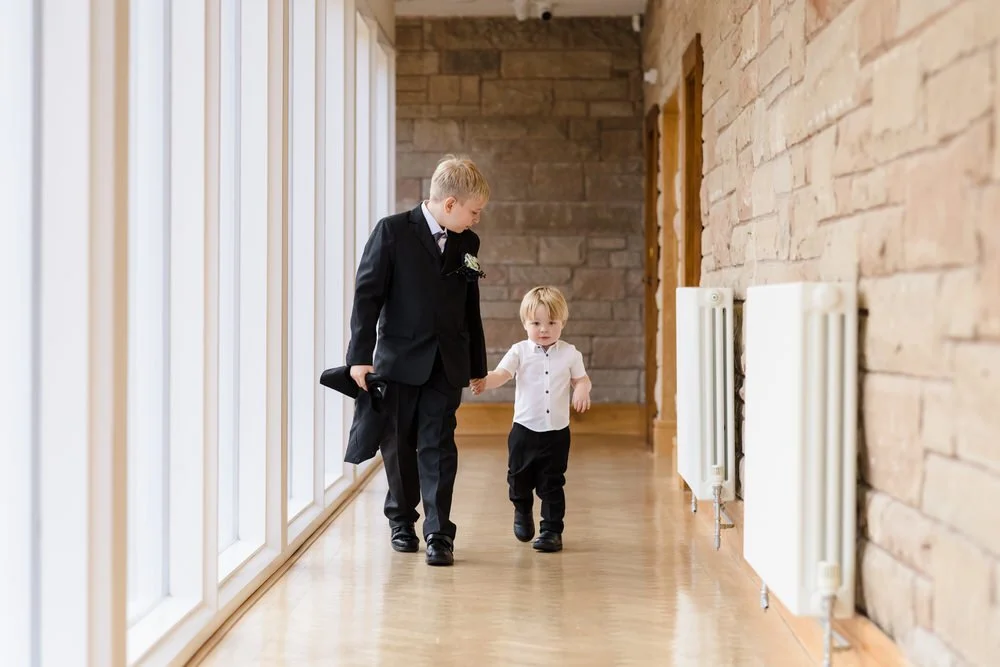 Page boy and young guest walking hand in hand through Arley House corridor, candid children photography by Once in a Lifetime Photography