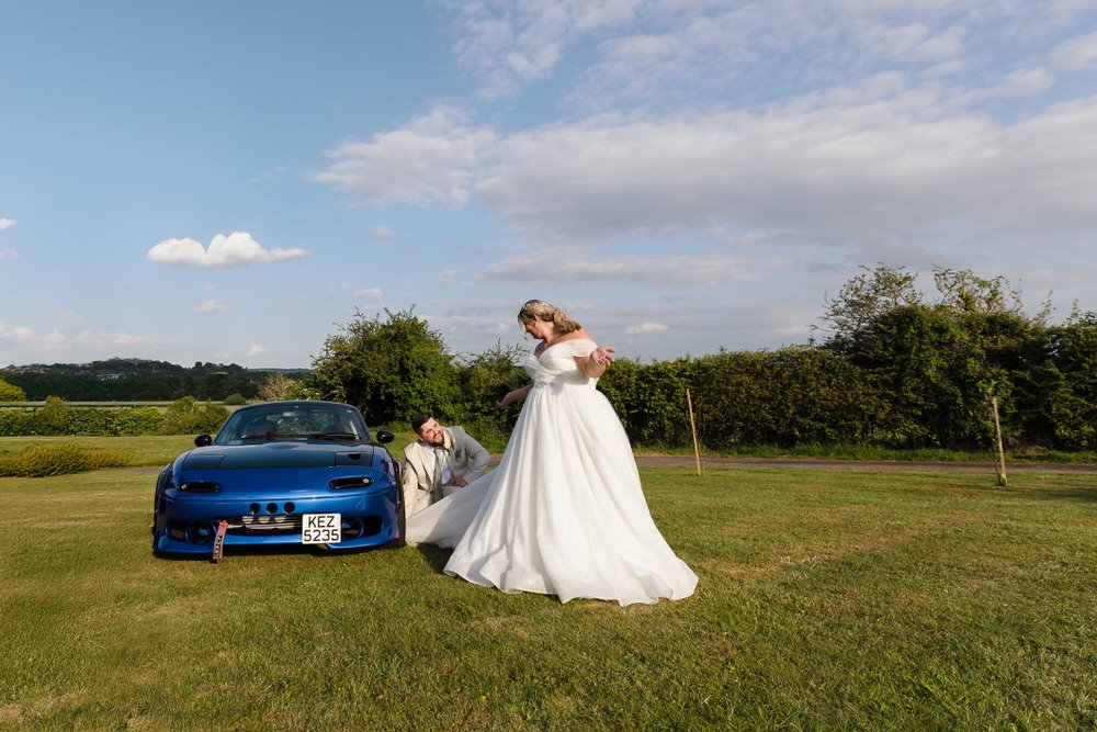 Bride in flowing white wedding dress and groom posing playfully with classic blue sports car at Bordesley Park Worcestershire wedding. Creative wedding photography capturing car enthusiast couple in countryside setting
