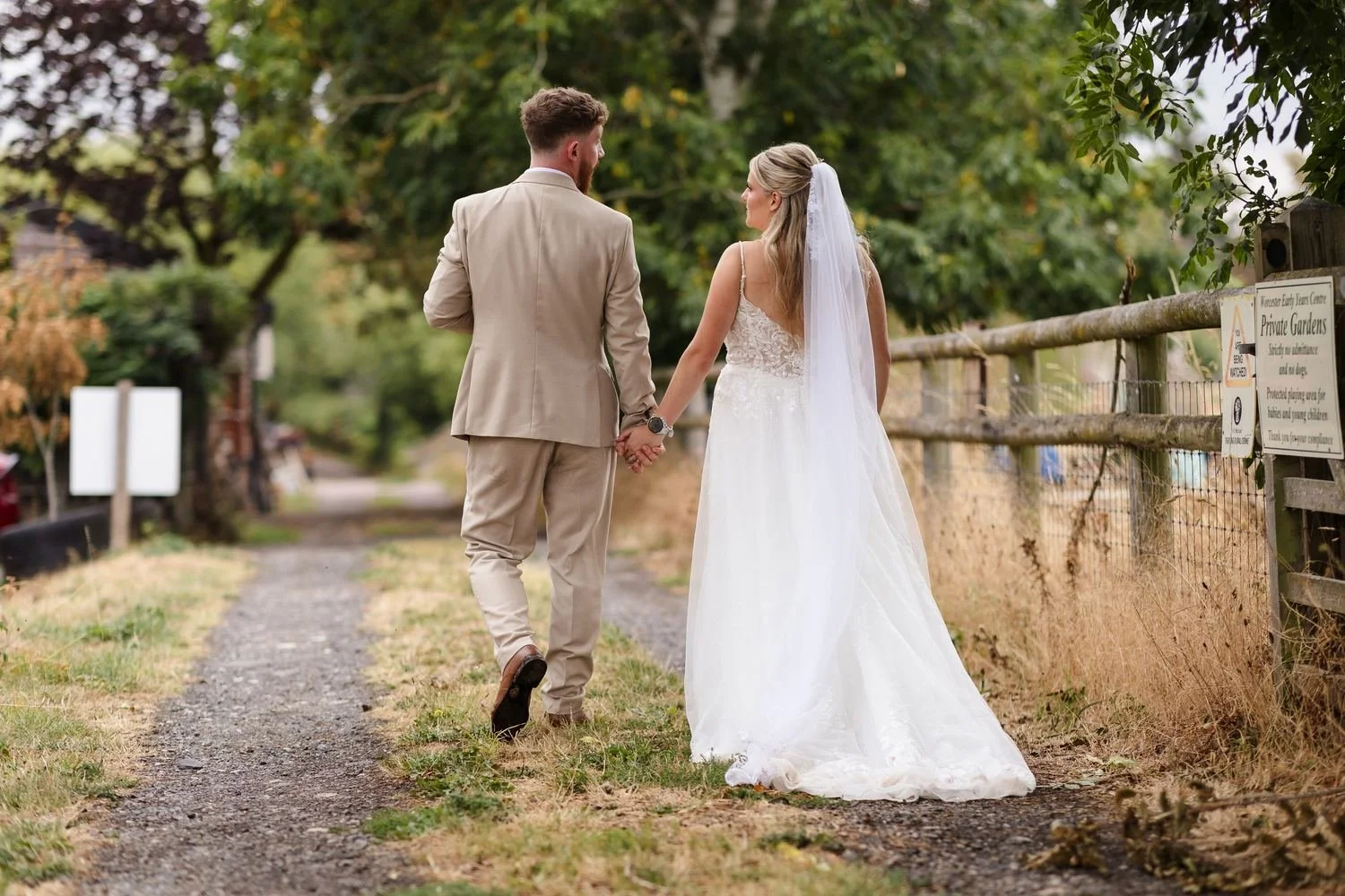 Abbie and Sean walking hand in hand along a gravel path beside a wooden fence, her long veil trailing behind
