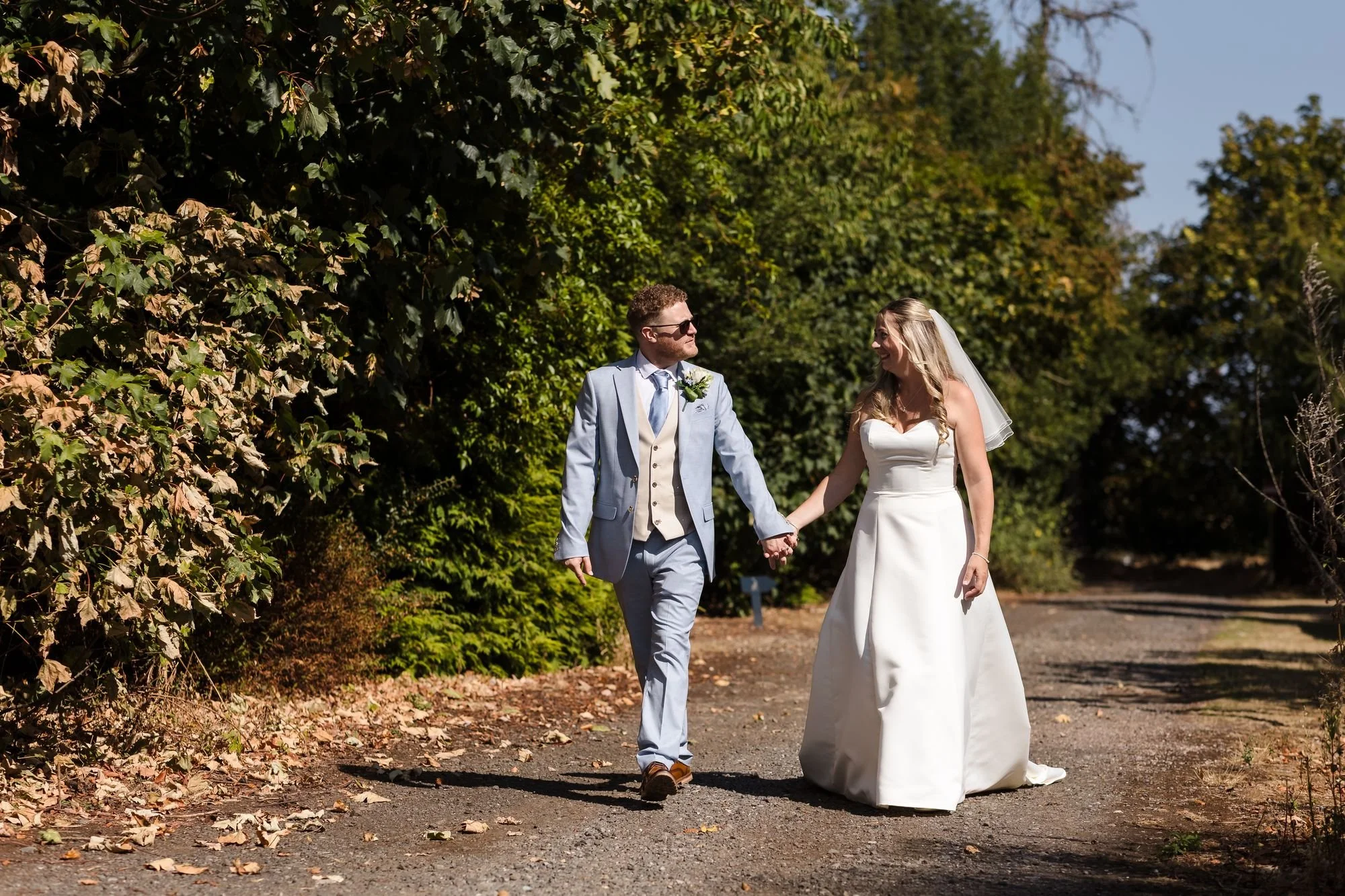 Cheryl and Josh walking hand in hand along a sunny tree-lined track in the grounds of Hogarths Stone Manor