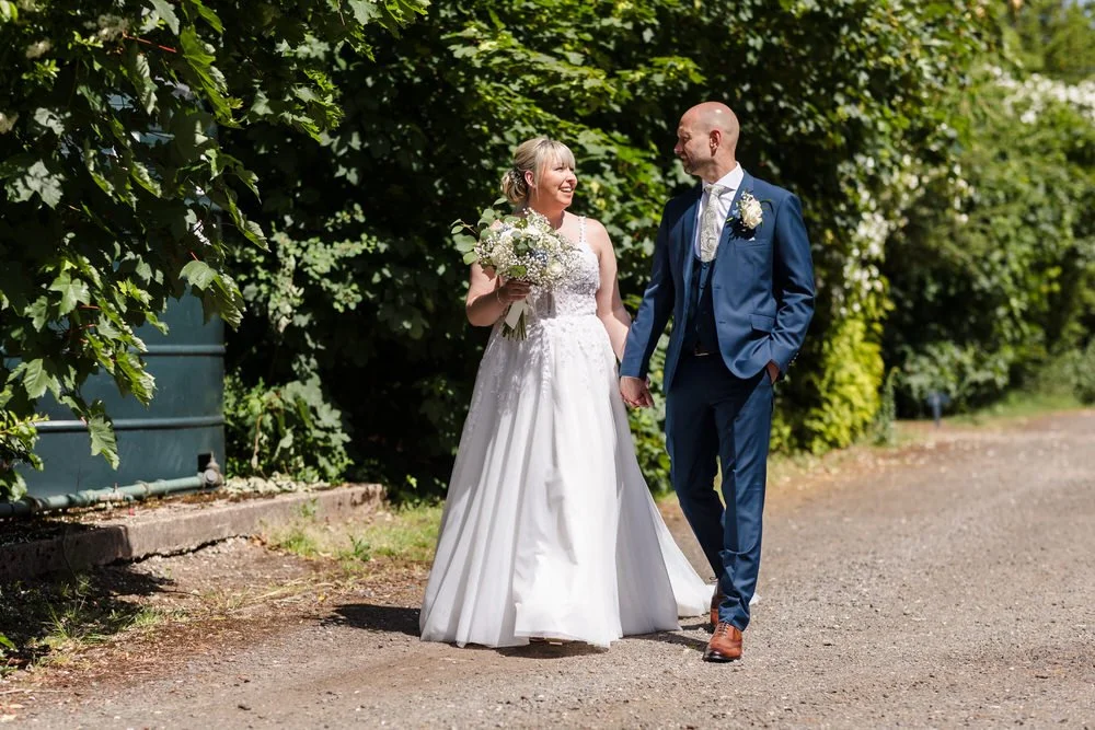 Bride and groom walking hand in hand along a tree-lined path at Hogarths Stone Manor