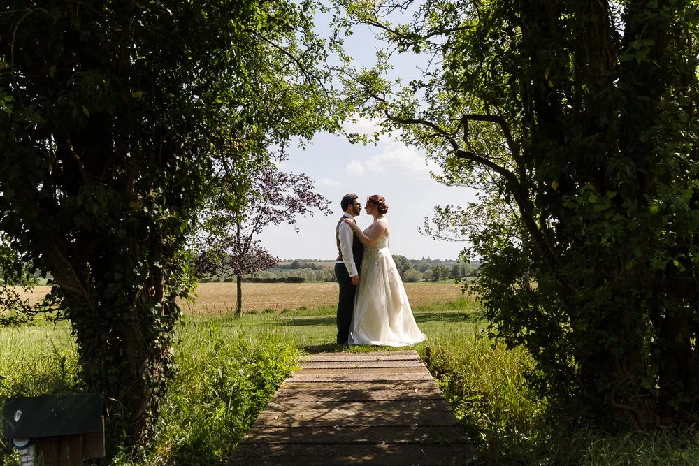 Bride and groom embracing under a tree archway at Huntsmill Farm, with the Buckinghamshire countryside beyond.