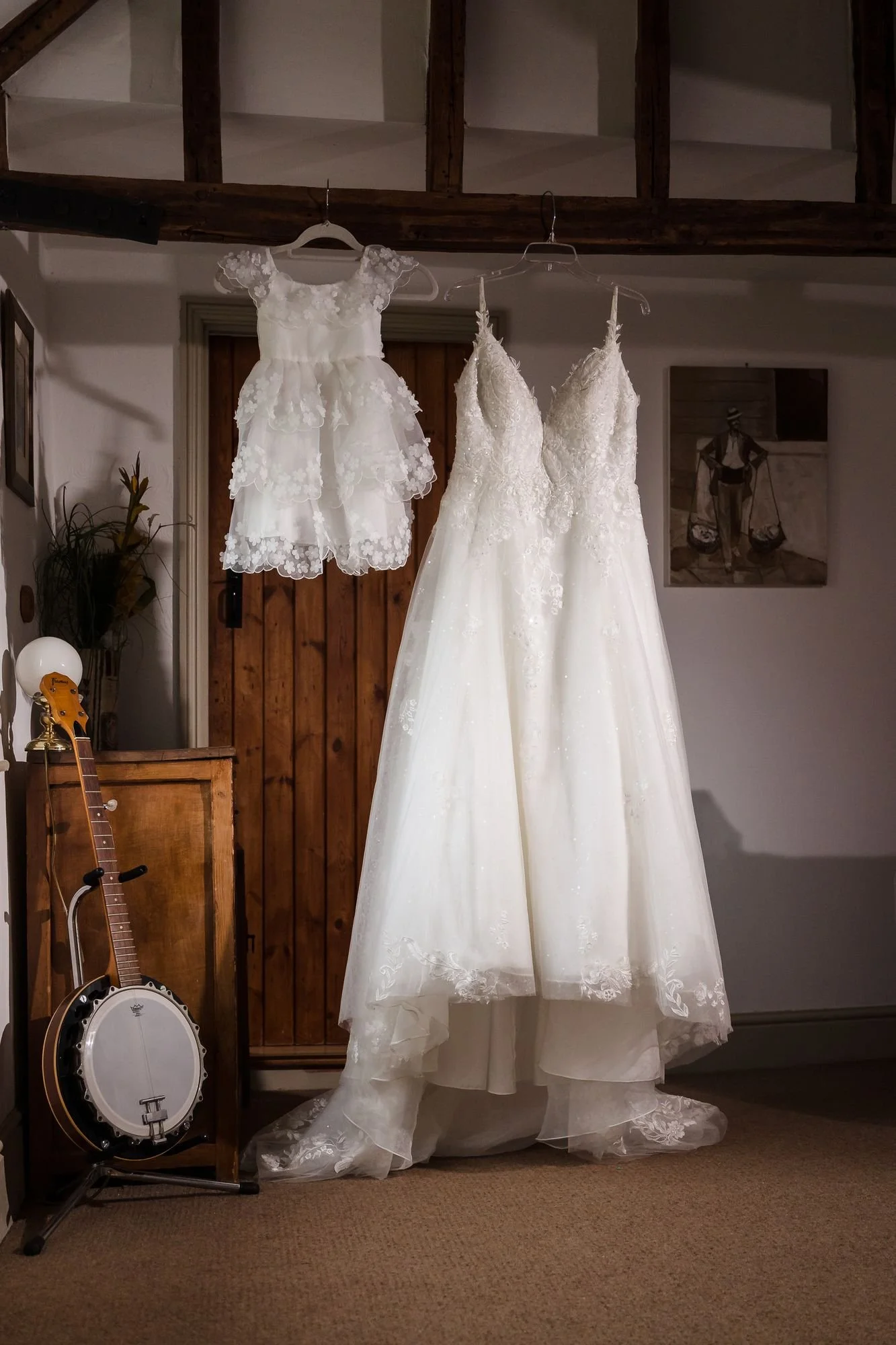 Abbie's lace wedding dress hanging from a beam beside a small lace flower girl dress, with a banjo in the corner of the cottage room