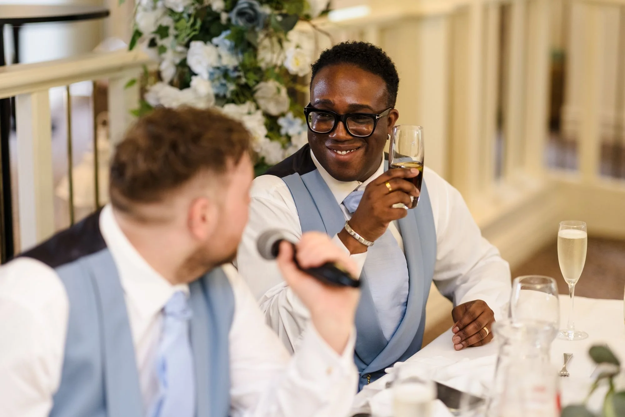 One groom raising a glass and smiling as his husband gives a speech into a microphone at the top table