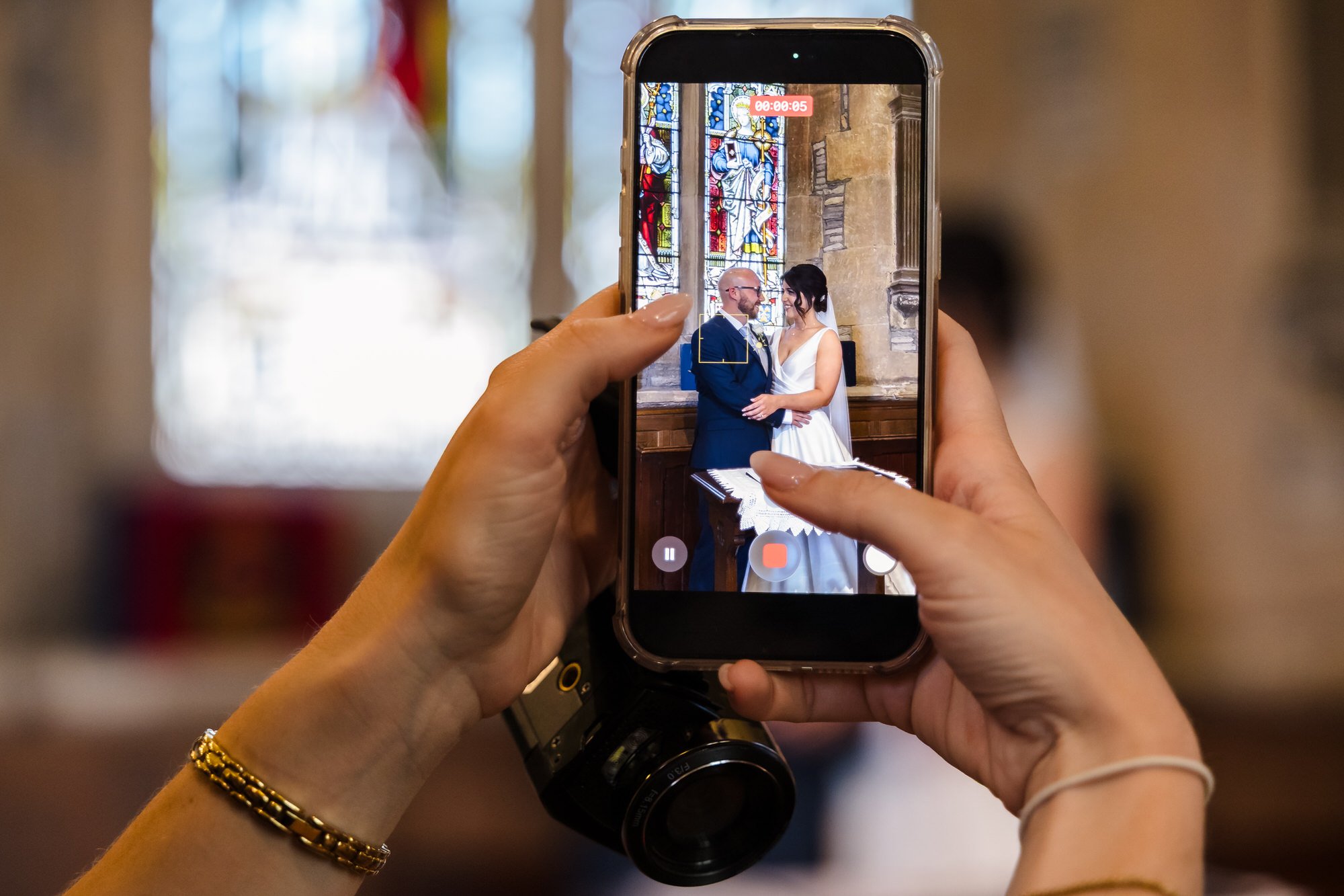 Guest capturing the ceremony on a phone alongside professional camera equipment, bride and groom visible on screen