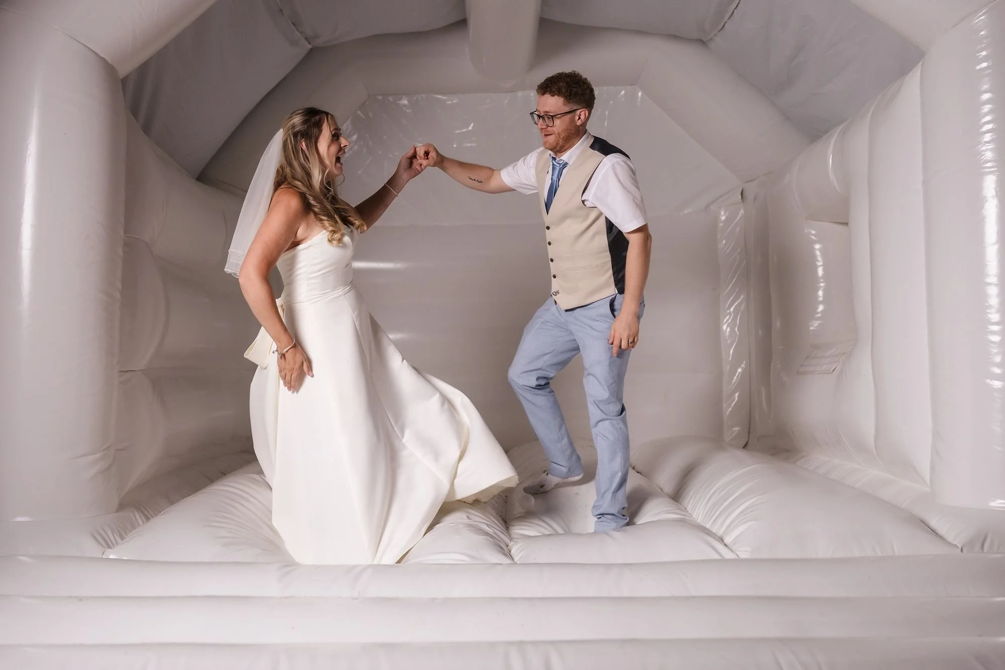 Cheryl and Josh bouncing hand in hand inside a white bouncy castle, lit with flash