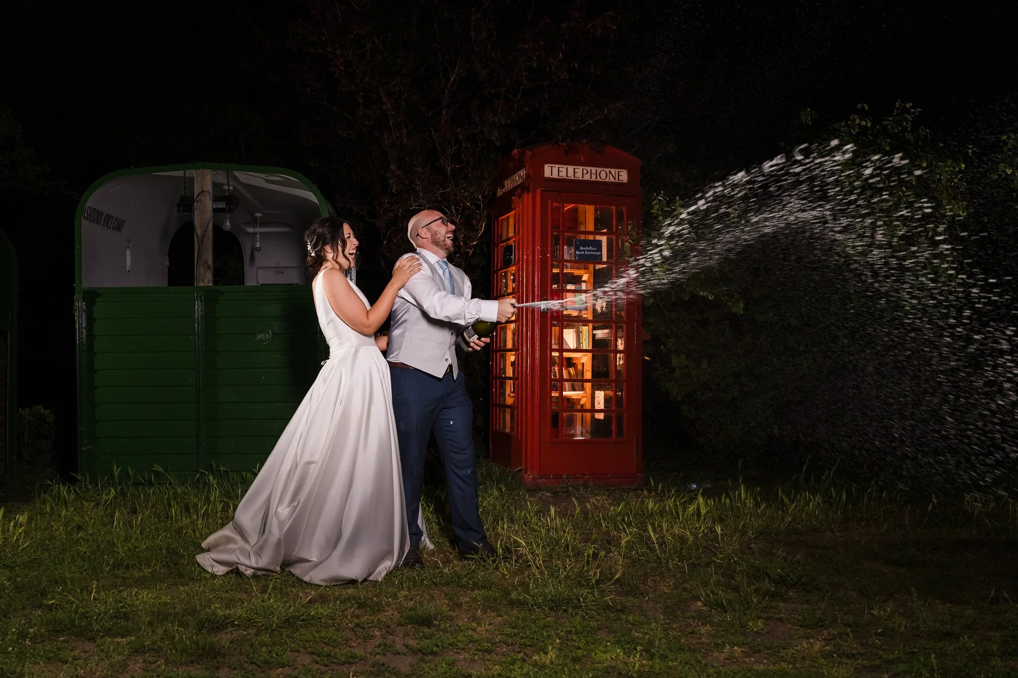 Groom spraying champagne beside a red telephone box, creative night portrait using off-camera flash at a Worcestershire wedding