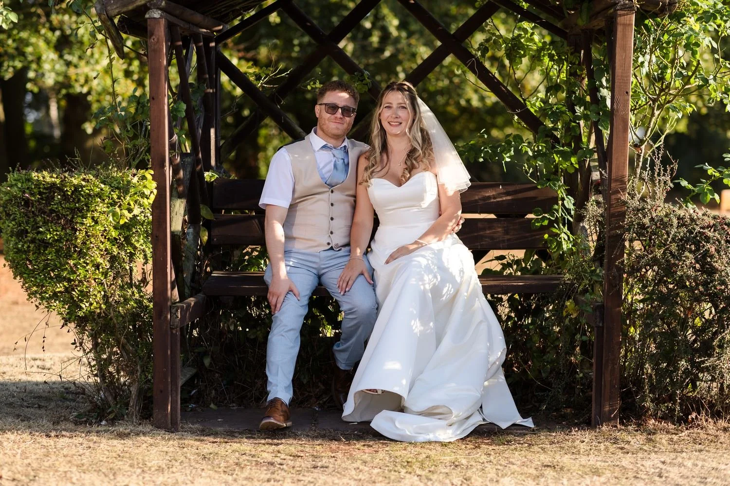 Cheryl and Josh smiling together on a wooden arbour seat in the gardens at Hogarths Stone Manor