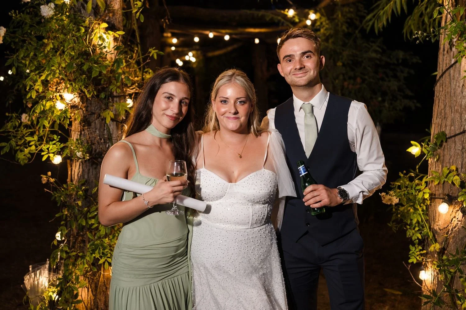 Three wedding guests smiling together under a fairy-lit pergola at night at Bennetts Willow Barn