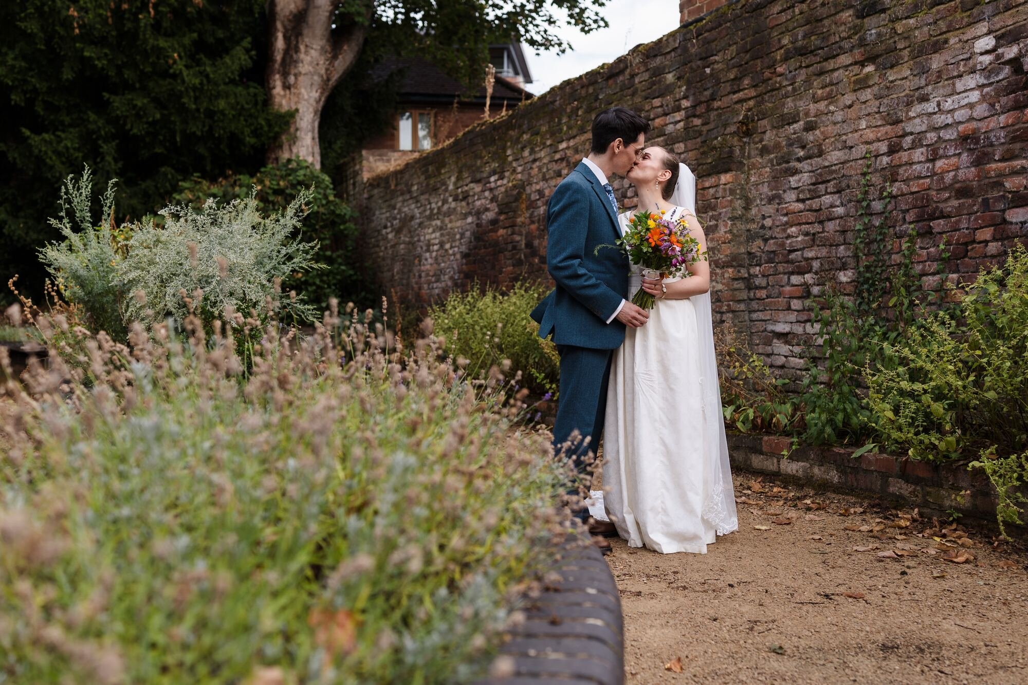 Isabelle and Chris kissing beside the flower border and old brick wall in the garden at The Commandery, Worcester, by Once in a Lifetime Photography