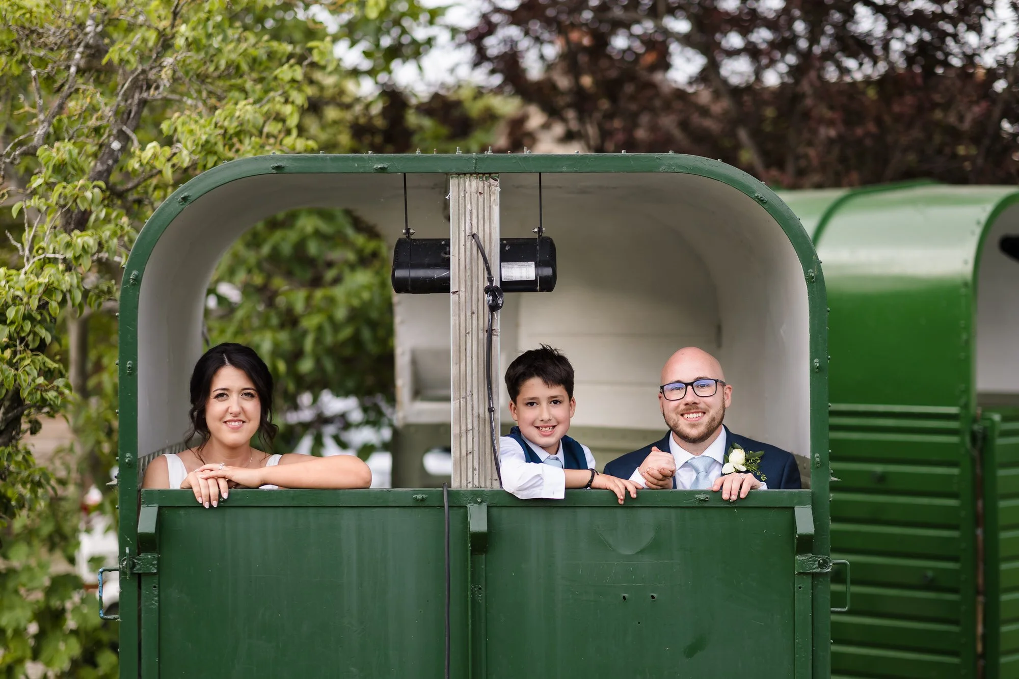 Bride, groom and page boy leaning out of a vintage horsebox photo booth at The Fleece Inn, Worcestershire wedding reception