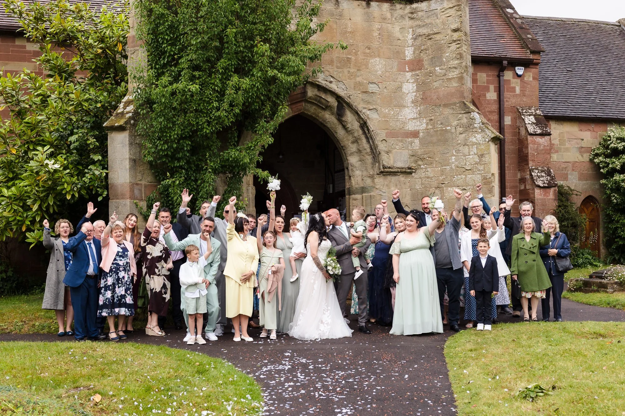 Full wedding party and guests cheering outside St Bartholomew's Church porch, Stourport-on-Severn.