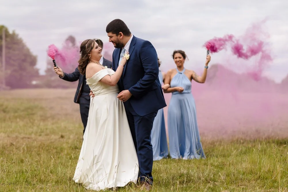 Couple share a dance in the field while bridesmaids create pink smoke for a dramatic portrait near Karma Salford Hall.