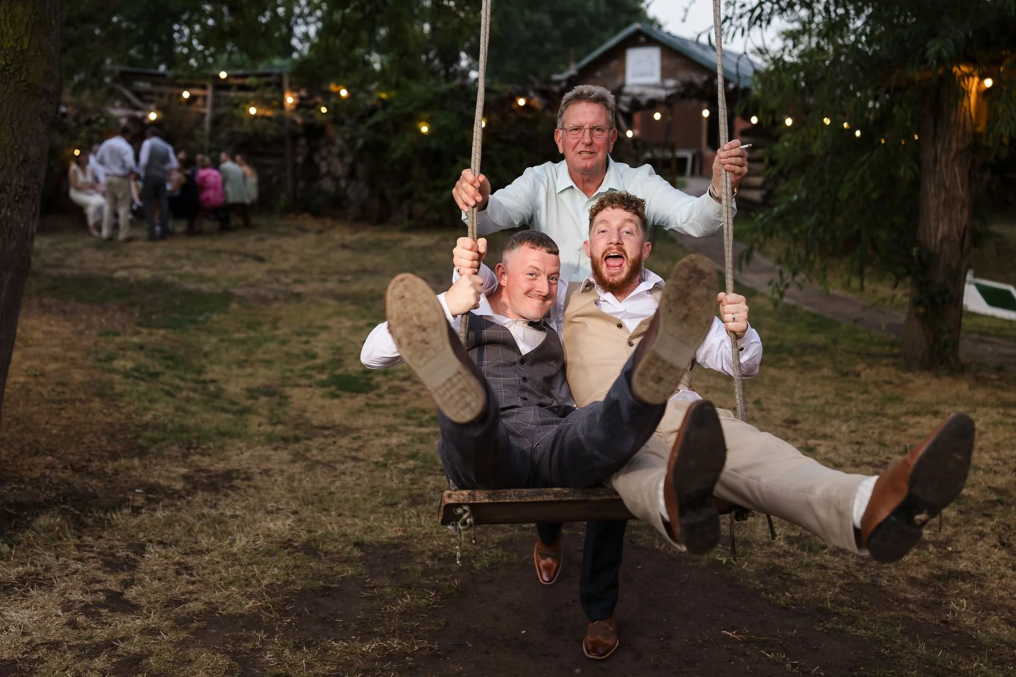 Sean and a friend kicking their legs up on the garden swing at dusk while a guest pushes them, festoon lights behind
