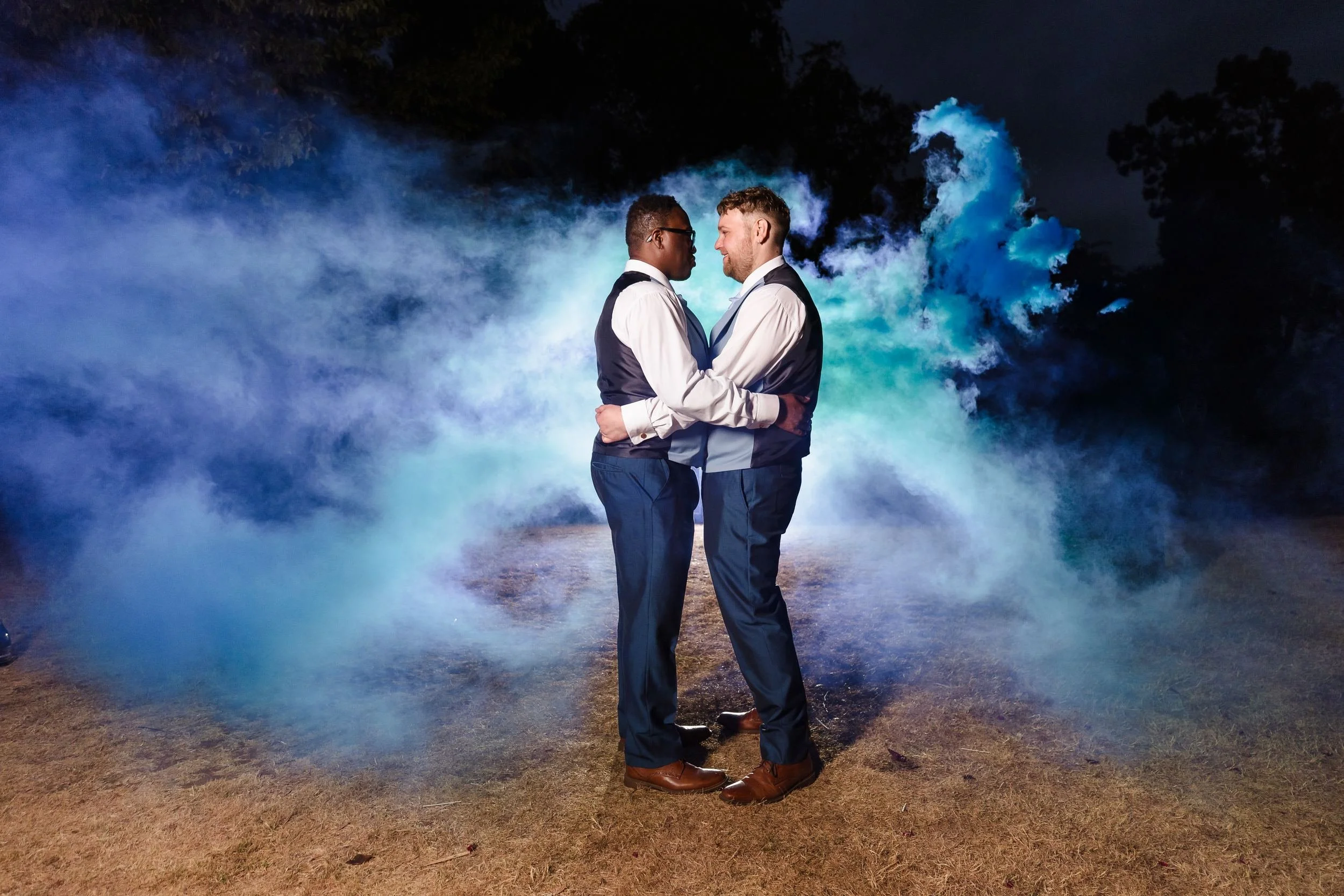 Liam and Shaun embracing at night, backlit by a flash through swirling blue and turquoise smoke