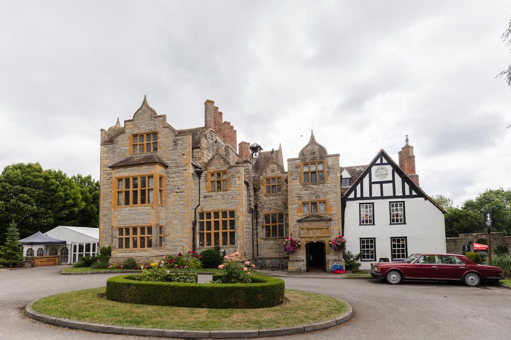 Front view of Karma Salford Hall’s stone and Tudor façades with a vintage red Rolls-Royce outside.