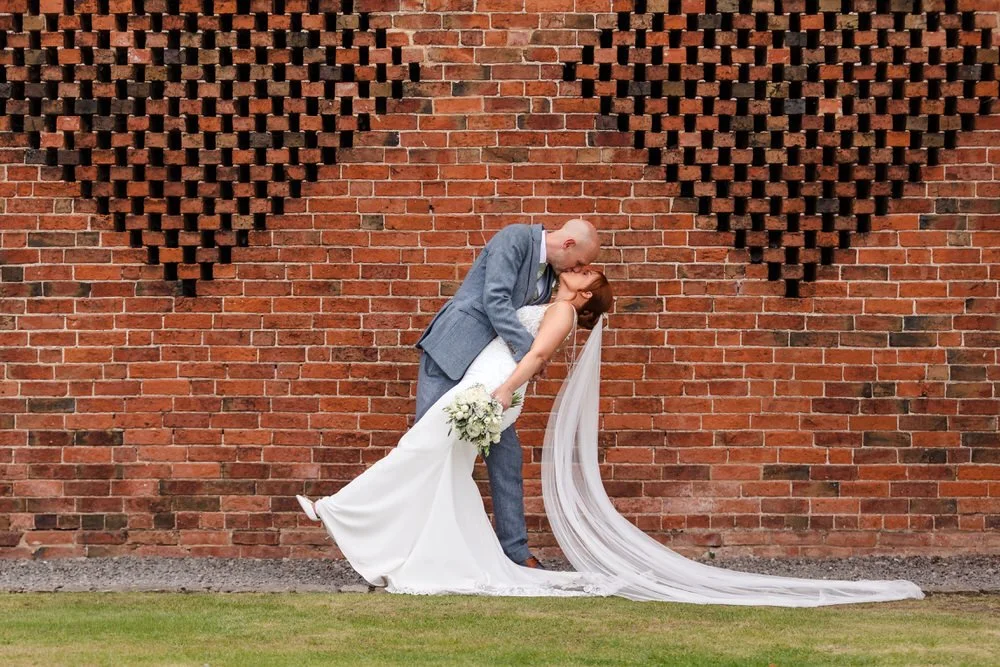 Groom dipping and kissing the bride against the iconic perforated brick wall at Shustoke Barn, bride's cathedral veil trailing across the lawn, holding a white rose bouquet in a fitted wedding gown