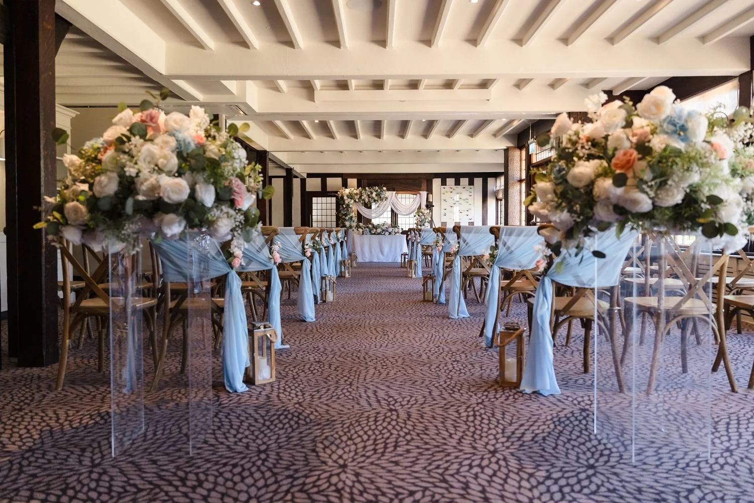 Tall floral pedestals and sky-blue drapes lining the aisle beneath the Drawing Room's timber beams